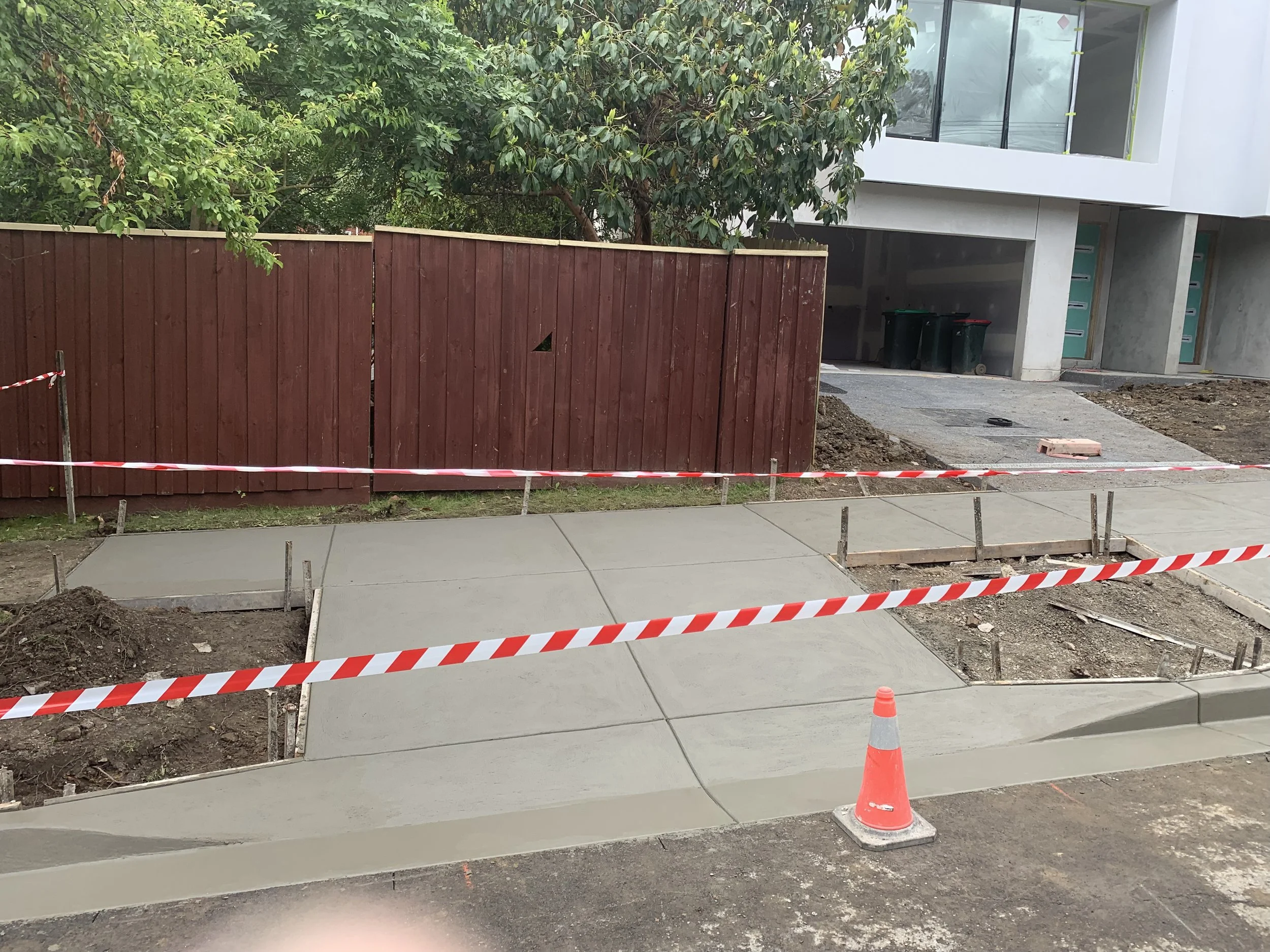 Sidewalk under construction with new concrete poured, surrounded by safety tape and orange traffic cone, in front of a modern building and red wooden fence.