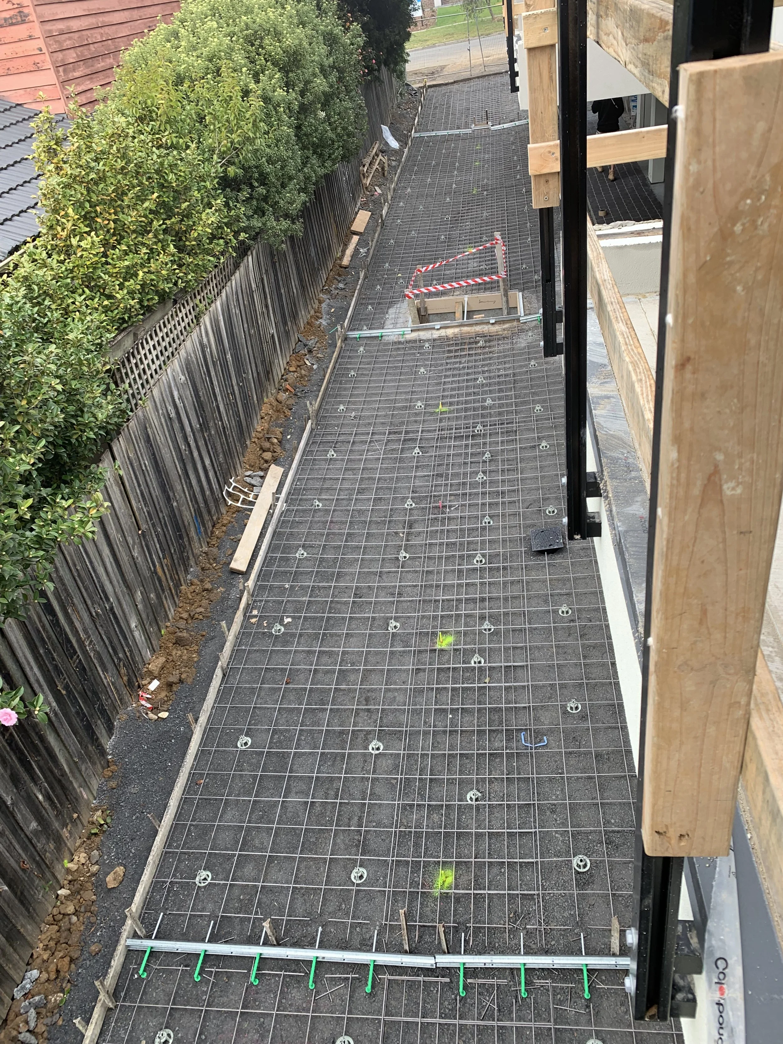 Construction site with metal grid flooring, wooden framing, and a proximity to a green hedge and wooden fence.