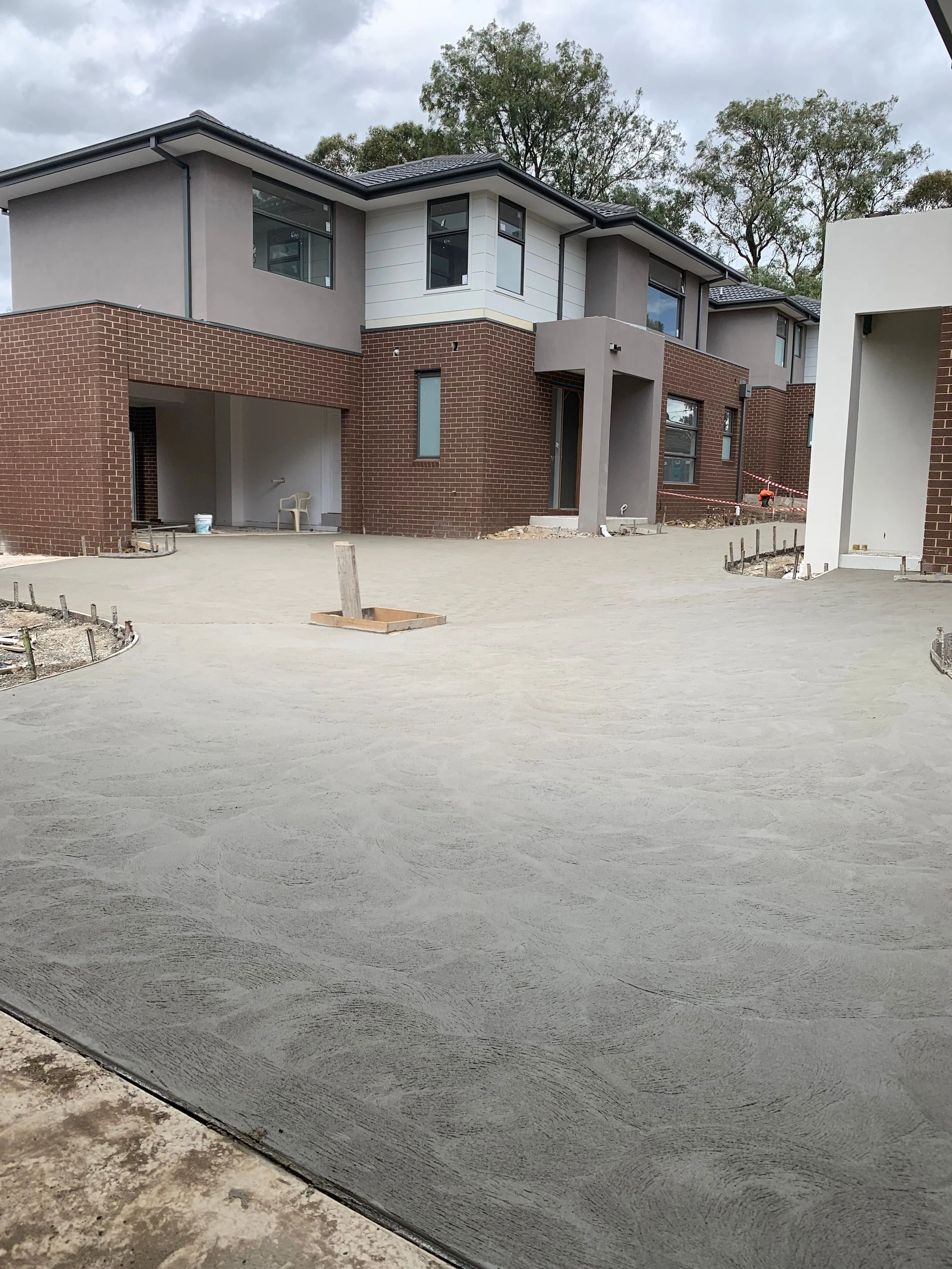 Newly constructed modern house with a concrete driveway in front, surrounded by trees, overcast sky, and construction materials nearby.