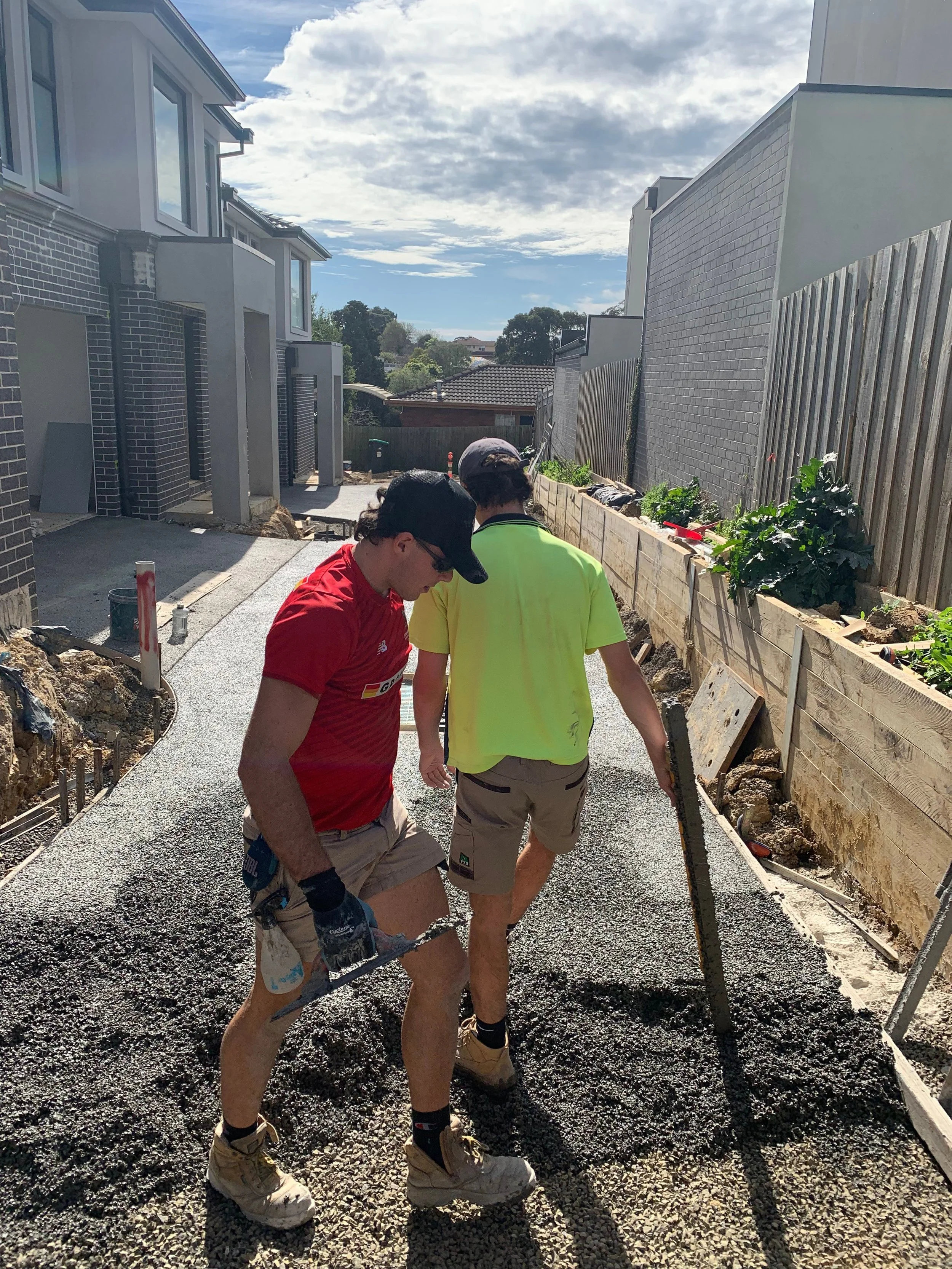 Two workers are laying gravel or concrete on a pathway in a residential back yard under a partly cloudy sky. One wears a red shirt, shorts, and gloves, holding a tool, while the other in a yellow shirt and shorts is standing nearby.