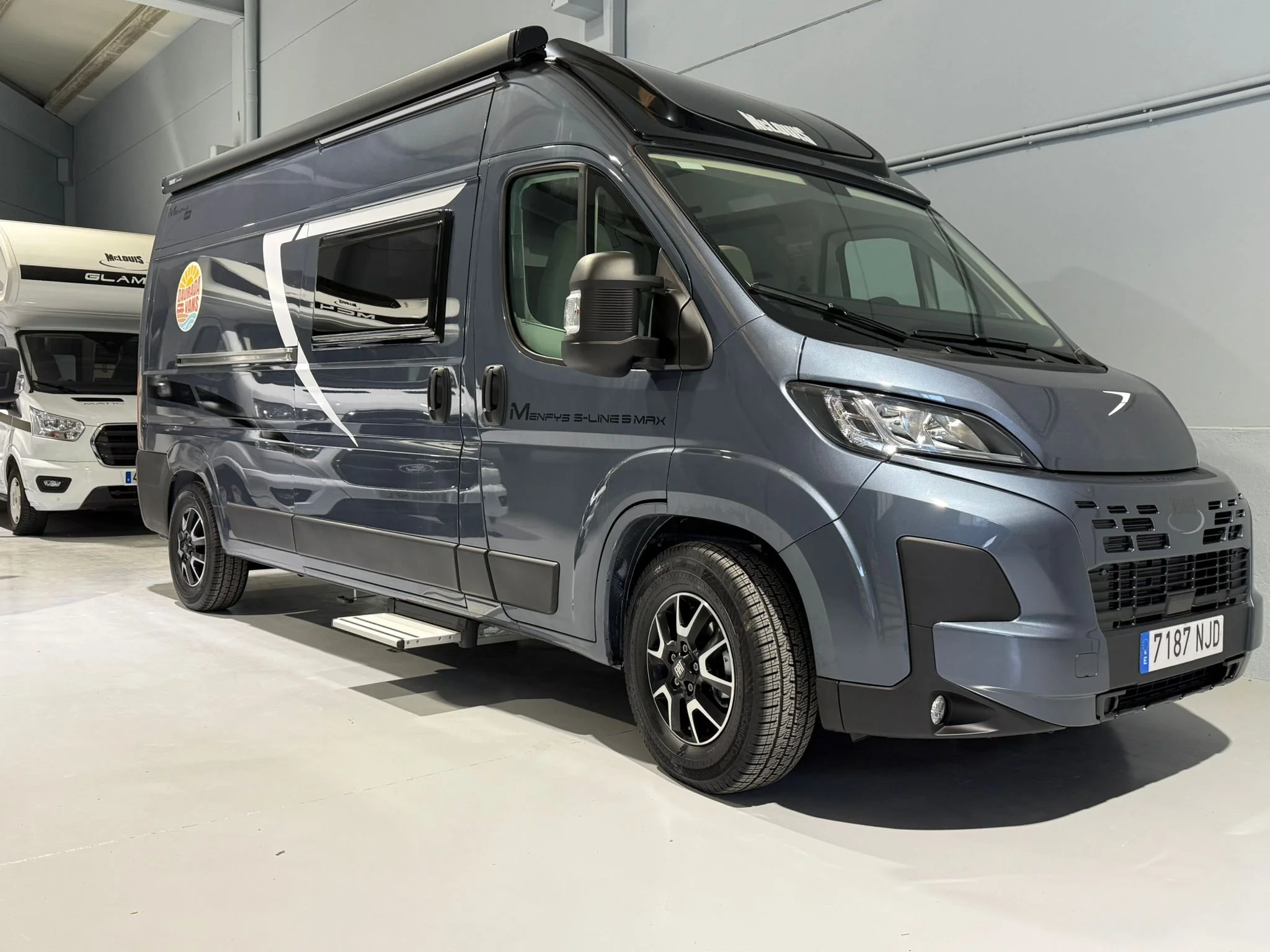 Furgoneta gris oscura con ventanas y detalles de diseño en negro, estacionada en un espacio interior con paredes grises y suelo claro.