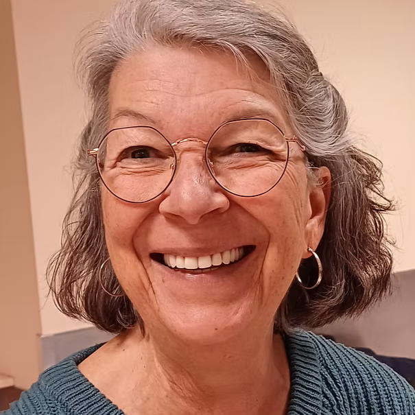 Une femme âgée souriante portant des lunettes rondes et des boucles d'oreilles, avec des cheveux gris tournés vers l'extérieur, portant un pull bleu.