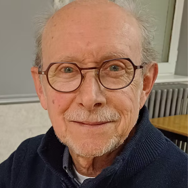 Un homme âgé souriant, portant des lunettes rondes, avec des cheveux gris clair, en intérieur.