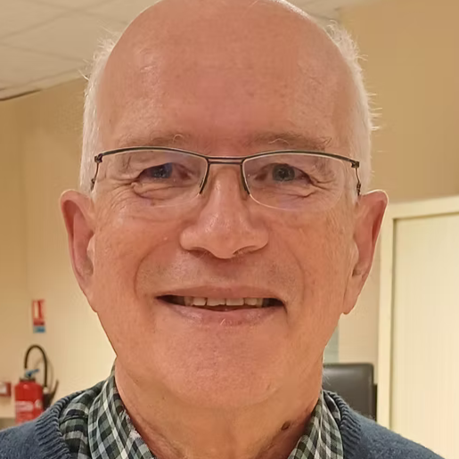 Un homme âgé souriant, portant des lunettes et une chemise à carreaux dans un bureau.
