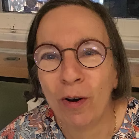 Une femme avec des lunettes rondes, cheveux bruns, portant un vêtement à motifs floraux, en intérieur.