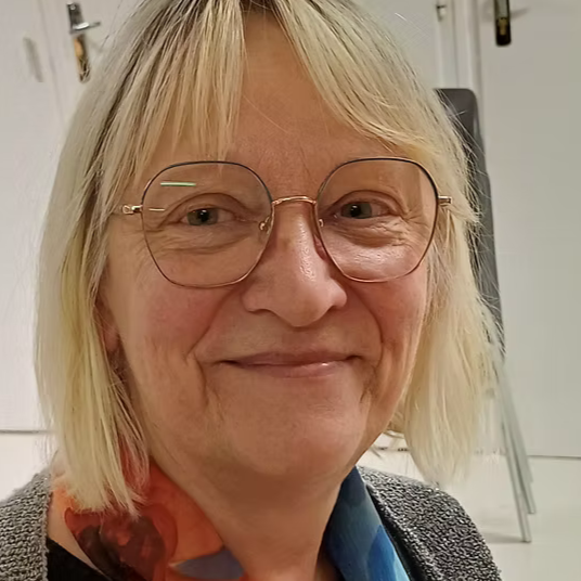 Un portrait d'une femme avec des lunettes rondes, cheveux blonds, souriante, dans un environnement intérieur.
