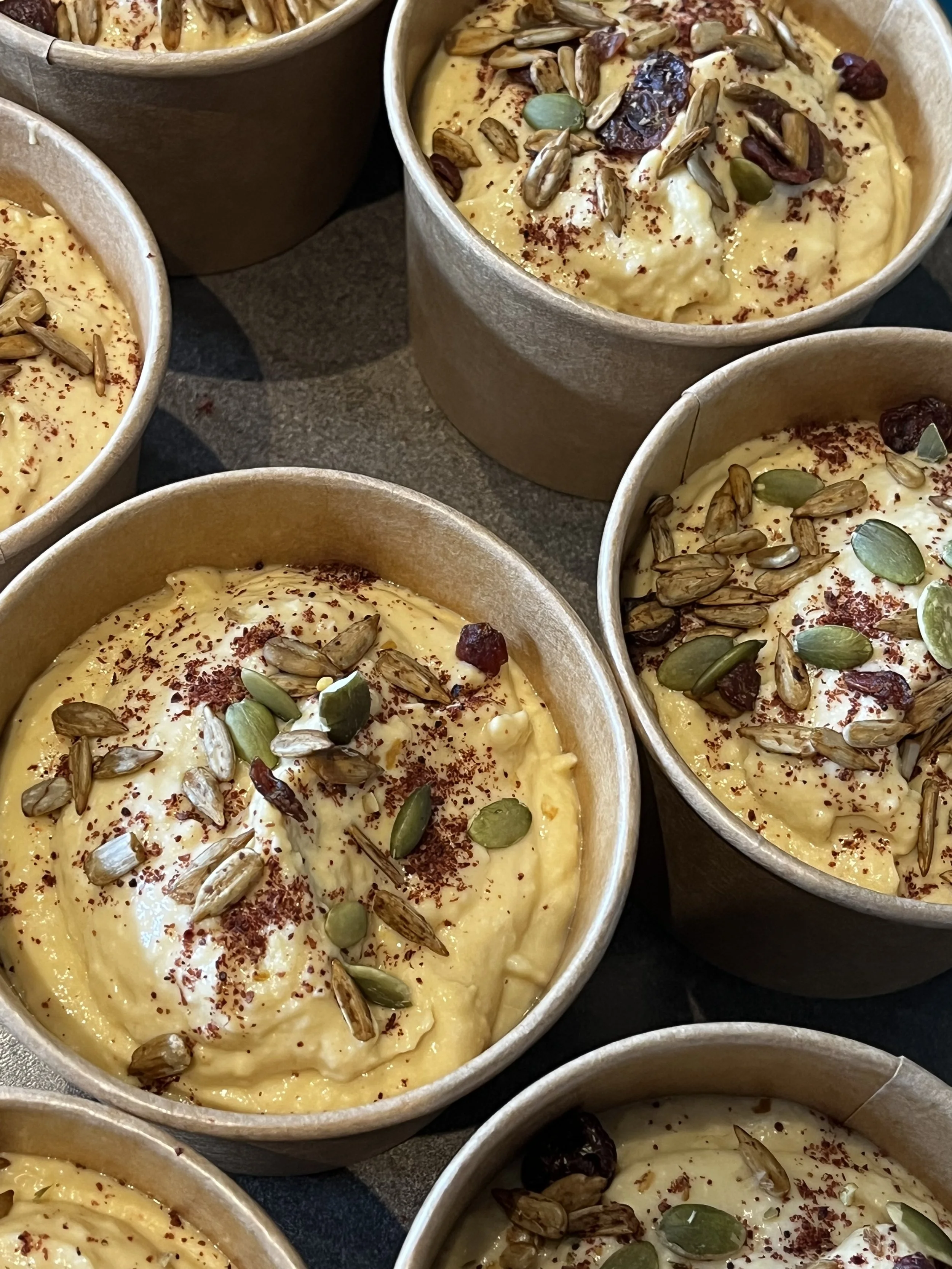 Creamy yellow dessert topped with pumpkin seeds, dried cranberries, and red spice powder in paper cups.