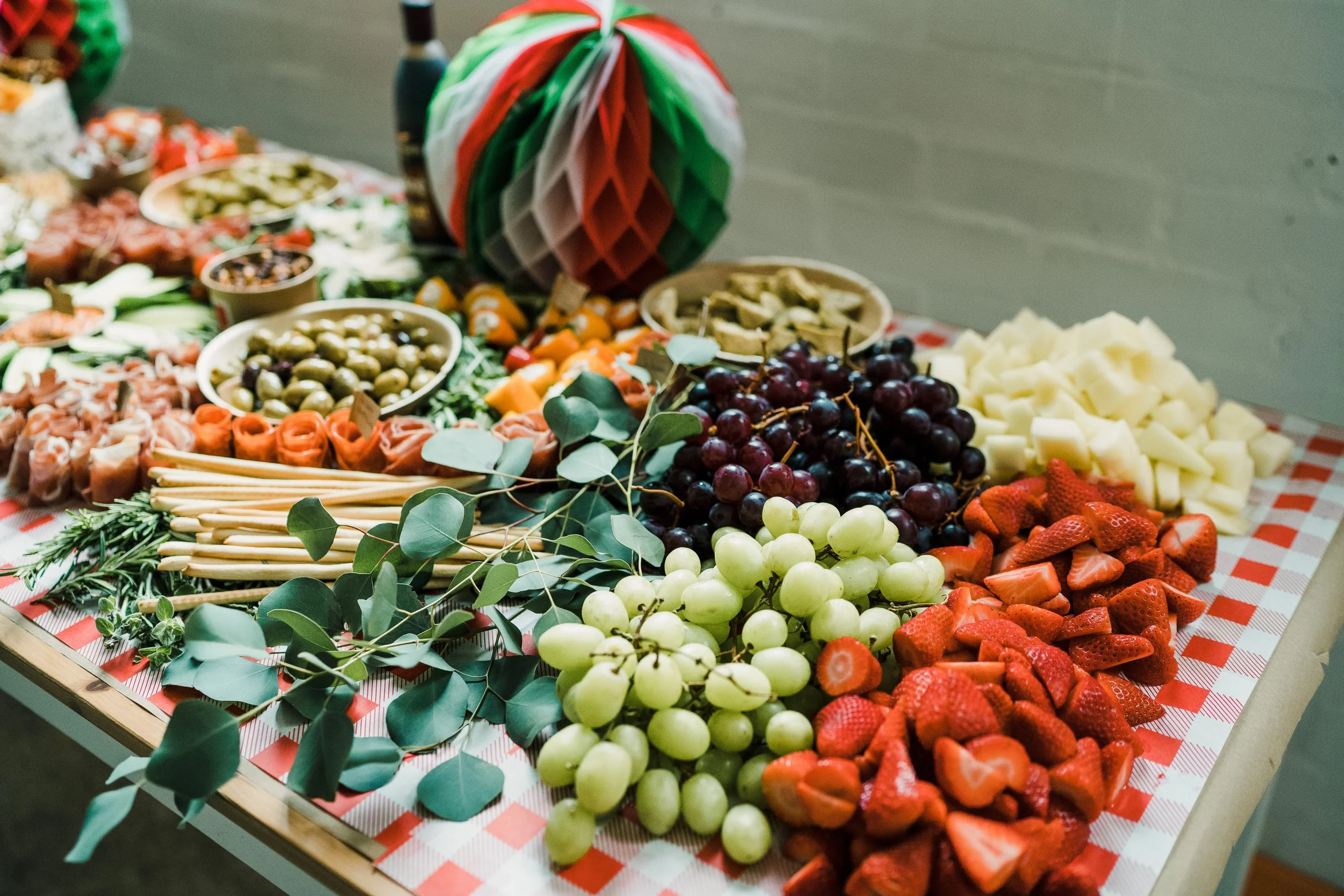 Colorful table display of assorted fruits, cheeses, and garnishes on a red and white checkered cloth with a fall-themed paper decoration in the background.