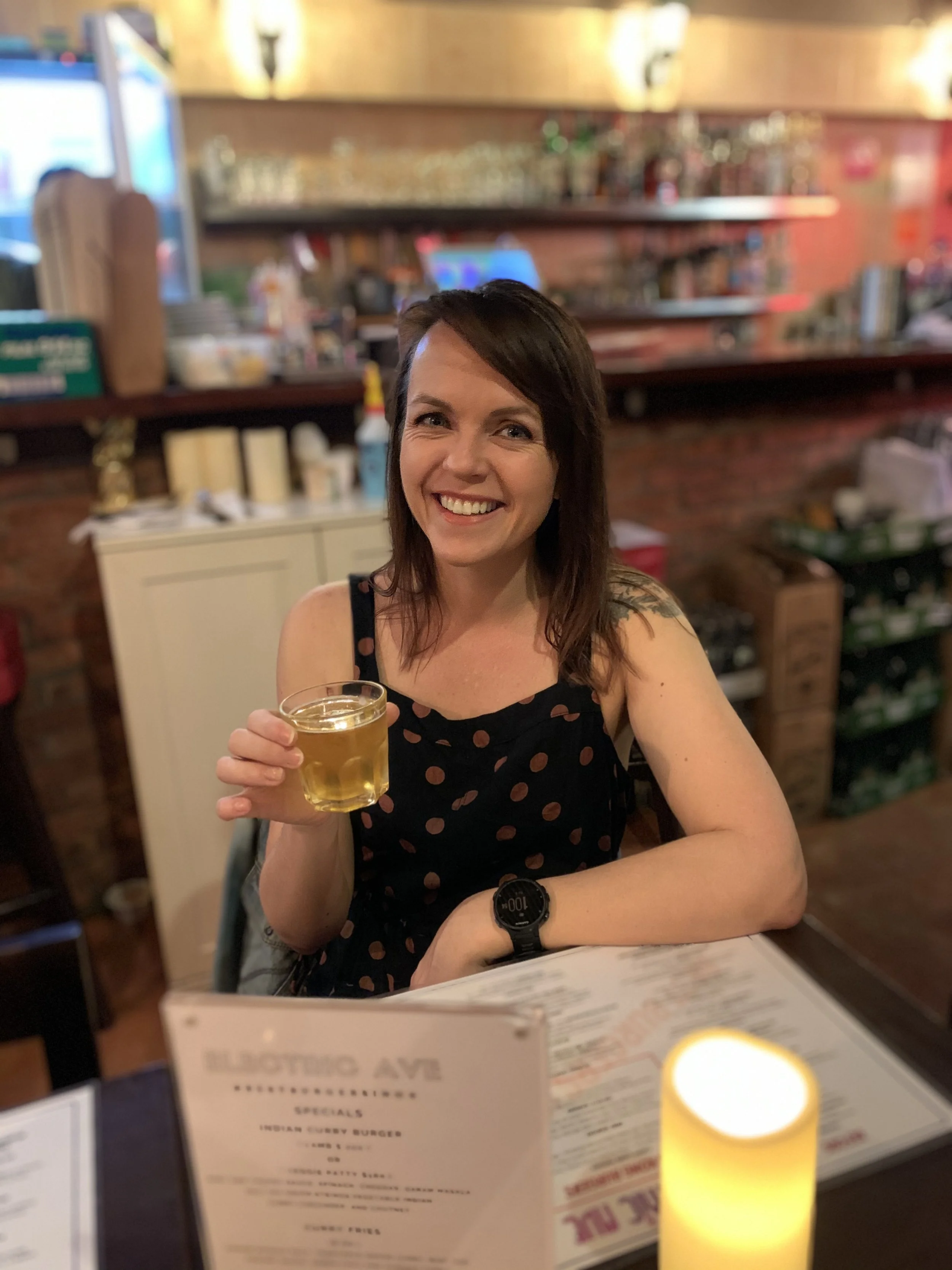 A smiling woman with dark brown hair holding a glass of beer at a restaurant or bar with shelves of bottles and glasses in the background.