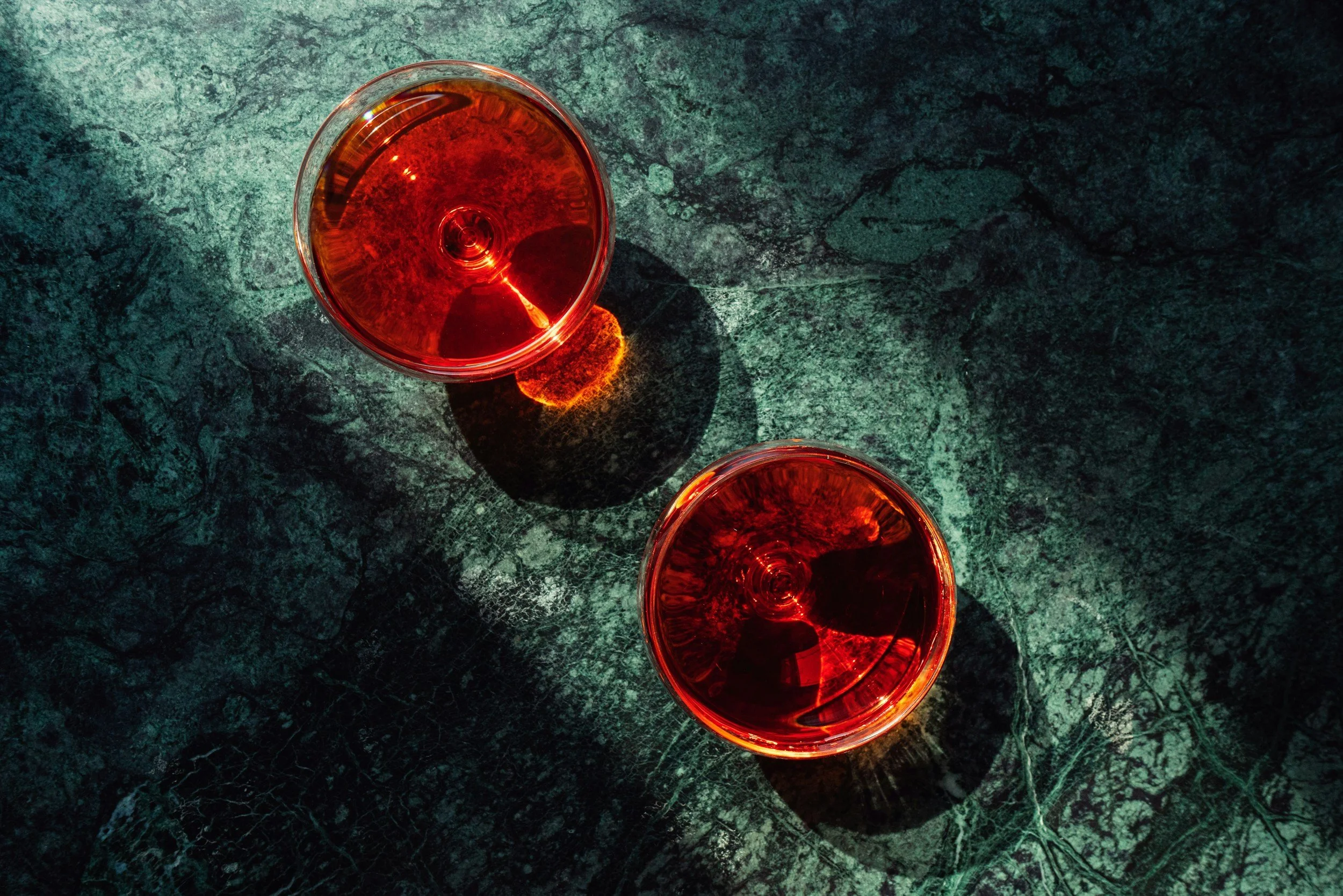 Two glasses of red wine on a dark green marble surface, casting shadows and reflections.
