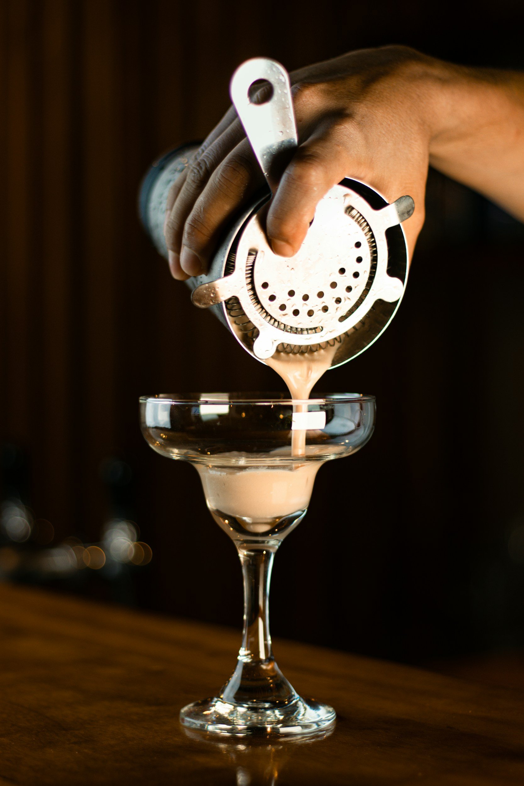 A bartender's hand pouring a creamy cocktail from a cocktail shaker into a glass on a wooden bar counter.