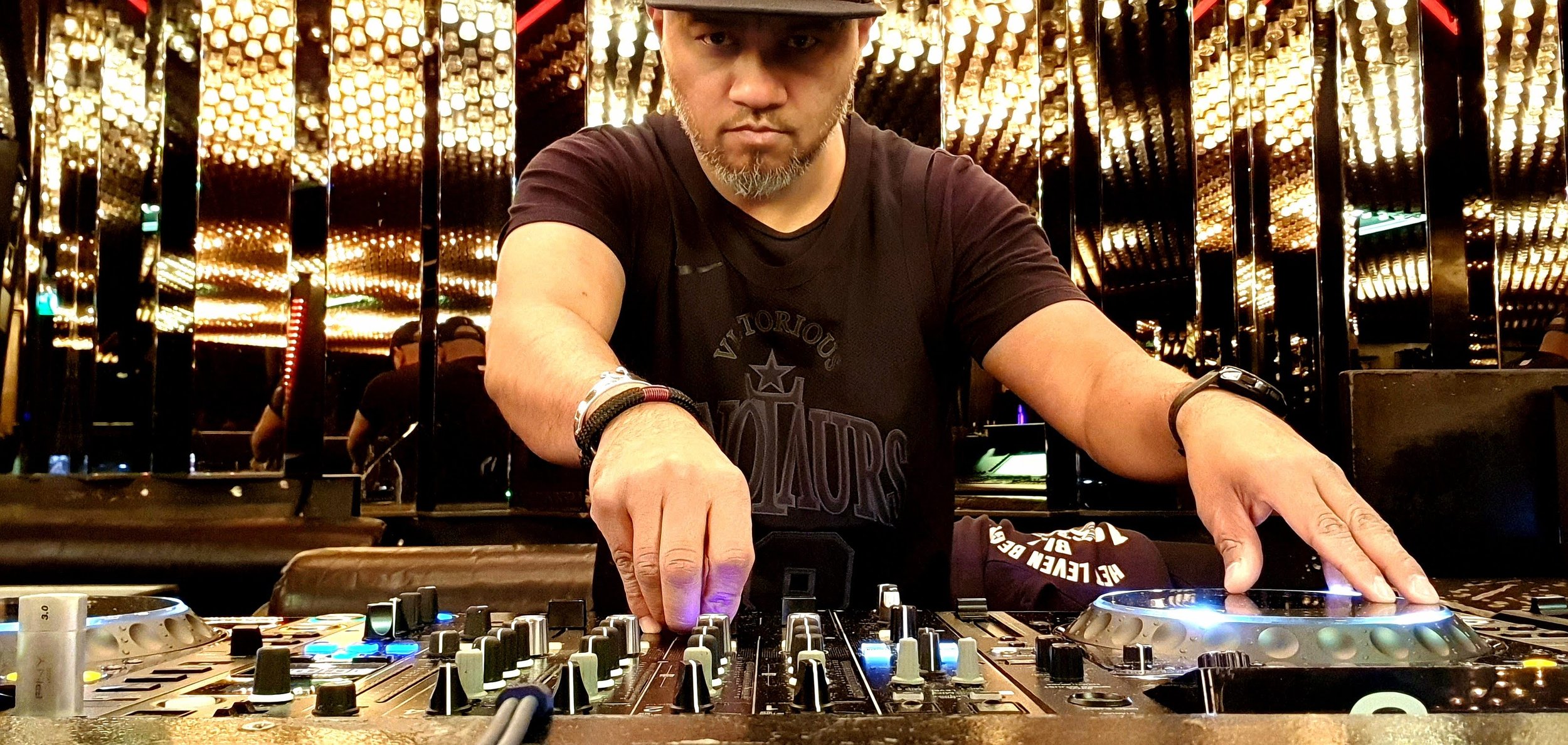 A DJ wearing a black T-shirt and cap is operating a mixing console in a venue with mirrored walls and warm lighting.