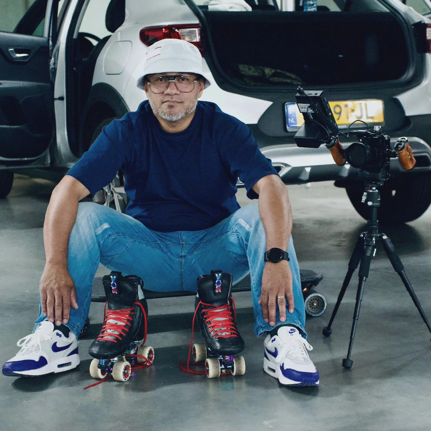 A man sitting on the floor in a parking garage, wearing athletic shoes, and roller skates, with a professional camera on a tripod beside him. He is dressed casually in a bucket hat, glasses, a blue t-shirt, and jeans. There are cars parked in the background.