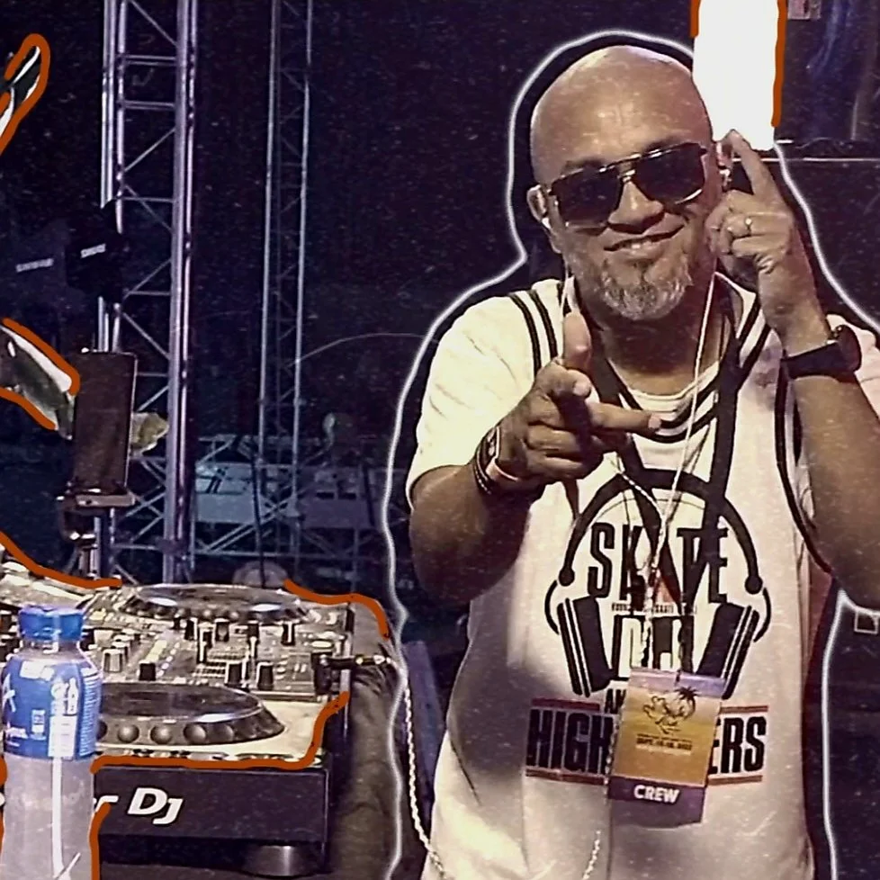 A bald man with a beard, wearing sunglasses, a white T-shirt with a graphic design, and a wristwatch, is smiling and pointing at the camera while holding a headphone with one hand. In the background, there is a DJ setup with turntables, a mixer, and a water bottle, at an event or concert.