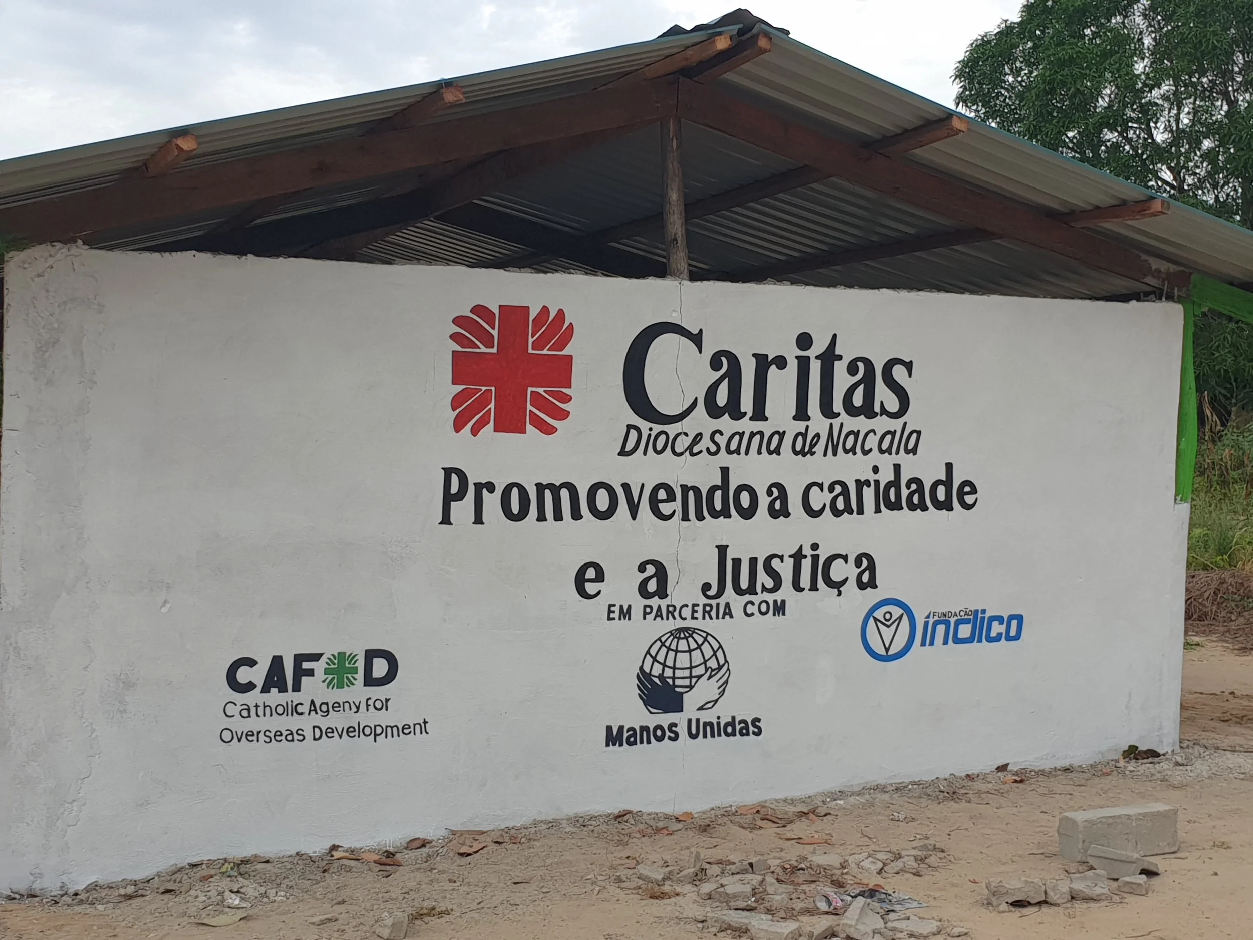 A painted white wall with a sign depicting a red cross with a stylized design, alongside text in Portuguese advertising Caritas diocese of Nacala, promoting charity and justice, with logos of Catholic Agency for Overseas Development (CAFOD), Manos Unidas, and Fundação Índico.