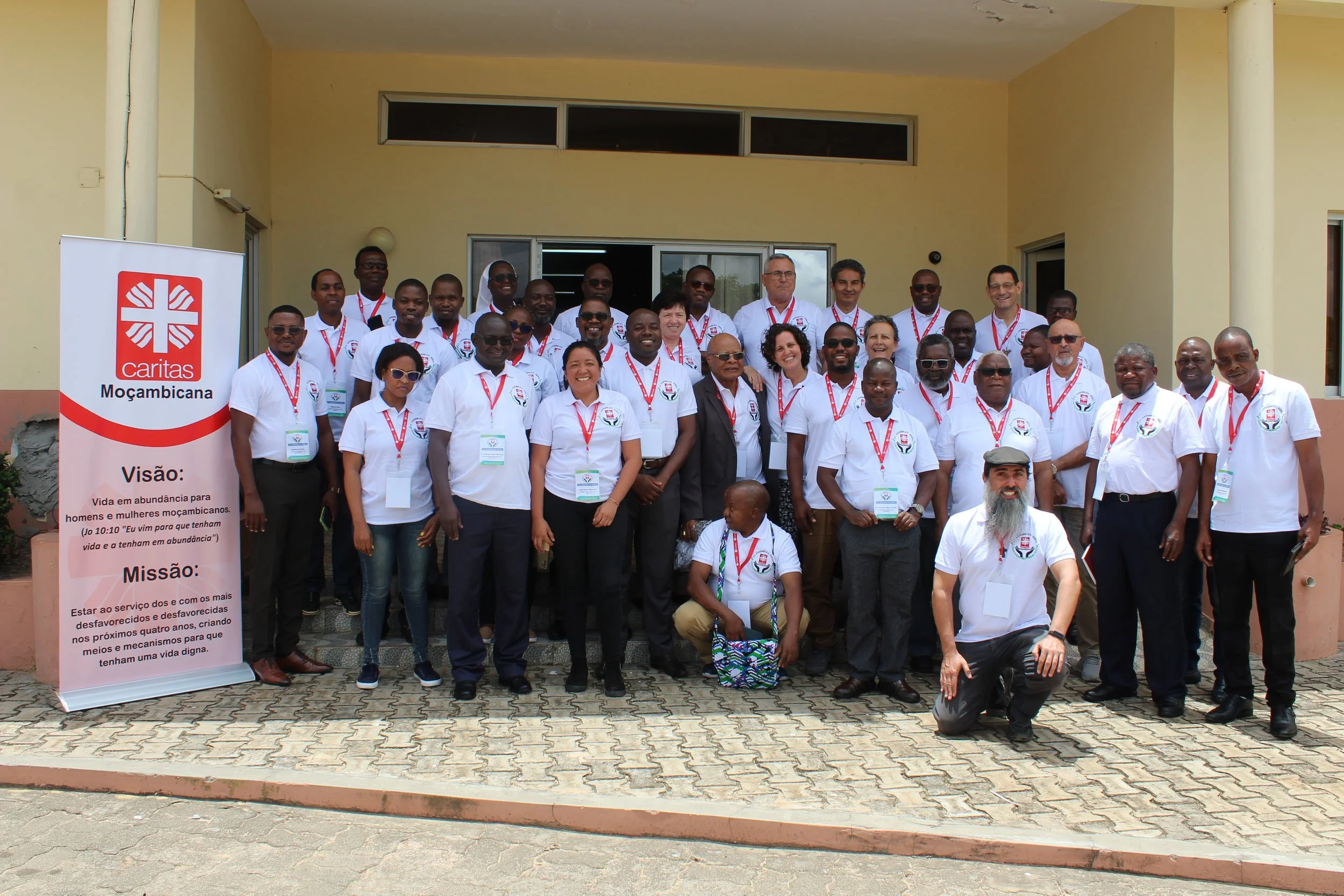 Group of people in white shirts posing for a photo outside a building with a "Caritas Moçambicana" banner on the left.