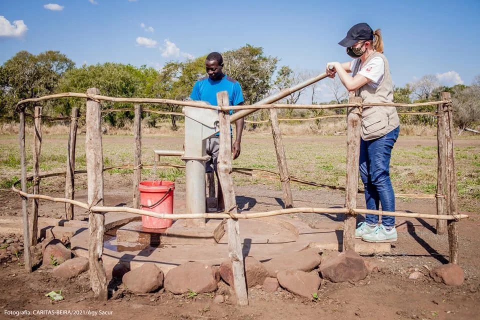 A woman and a man are using a hand water pump outdoors, surrounded by a small wooden fence, with trees and a field in the background.