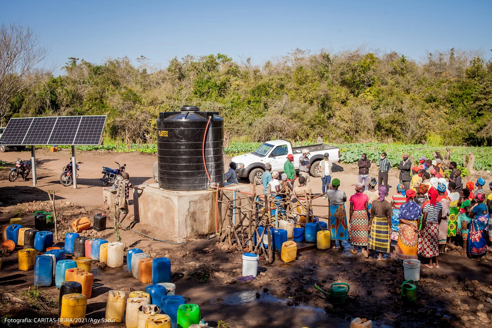A group of people, mostly women and children, gather outdoors near a large black water tank with orange hoses, in a rural setting with trees in the background. Several colorful water containers and buckets are on the ground, and a few motorcycles and a white pickup truck are parked nearby. Solar panels are installed on the left side of the image.