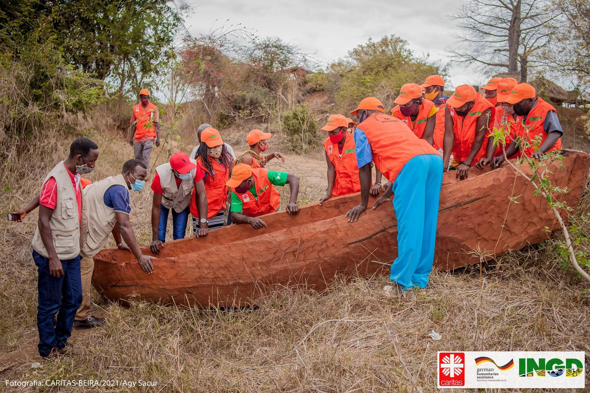 Group of people, including several wearing orange vests, orange hats, and face masks, lifting a large, carved wooden canoe or boat in an outdoor setting with trees and dry grass.