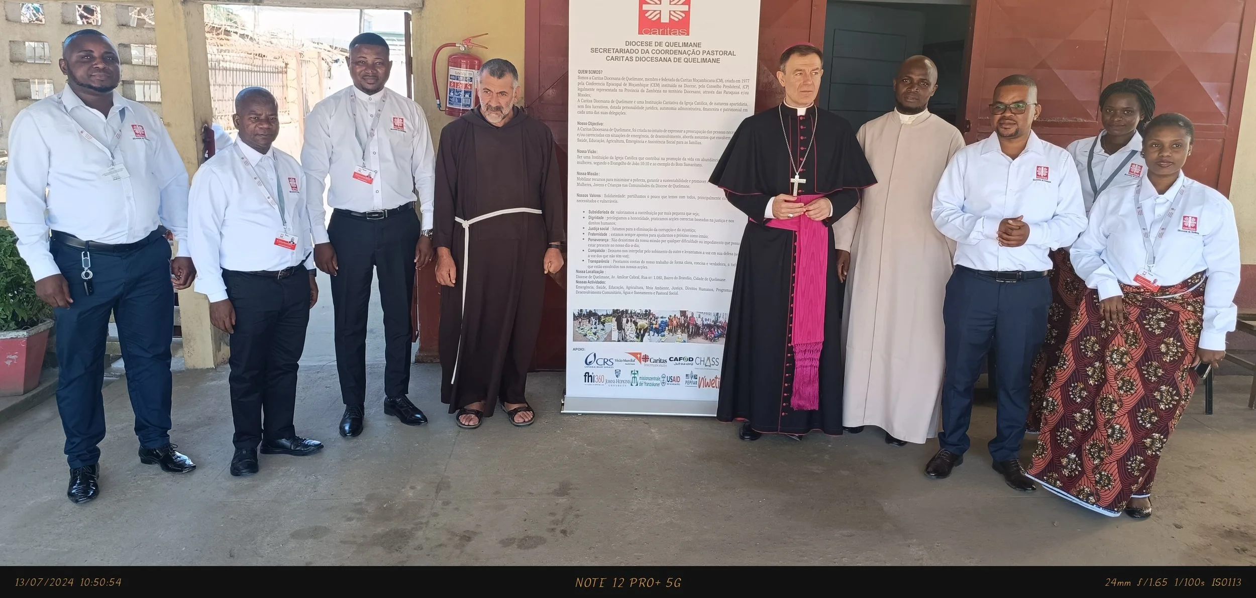 Group of nine people standing indoors, including religious and community leaders, in front of a poster with text and logos, wearing formal and religious attire.