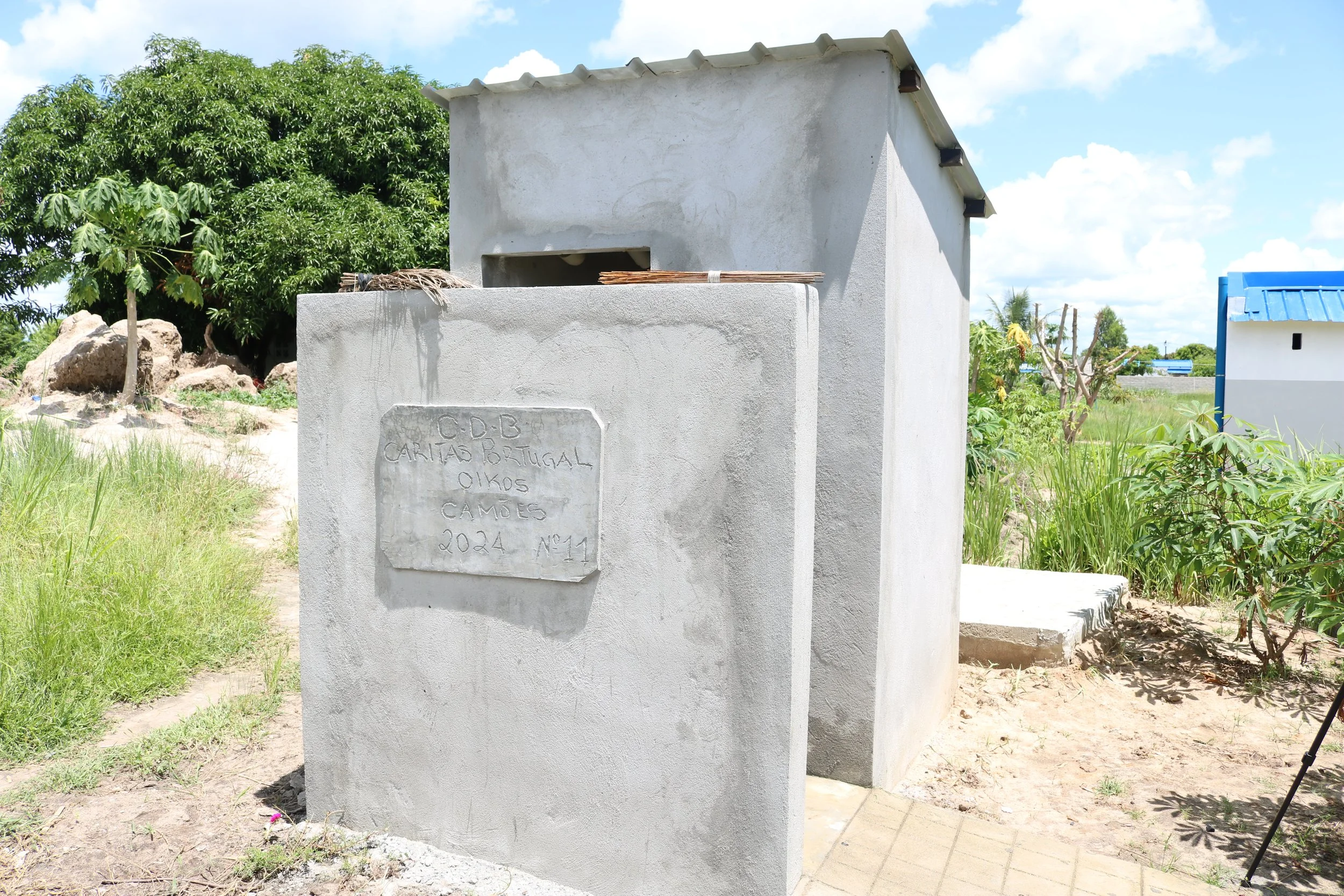 Small gray concrete structure with a plaque on the front, located outdoors in a rural area. The plaque reads 'C.C.D.B. Caritas Portugal 01 KOS Campos 2024 No 11'. Surrounding the structure are green trees, grass, and other small buildings, with a bright blue sky overhead.