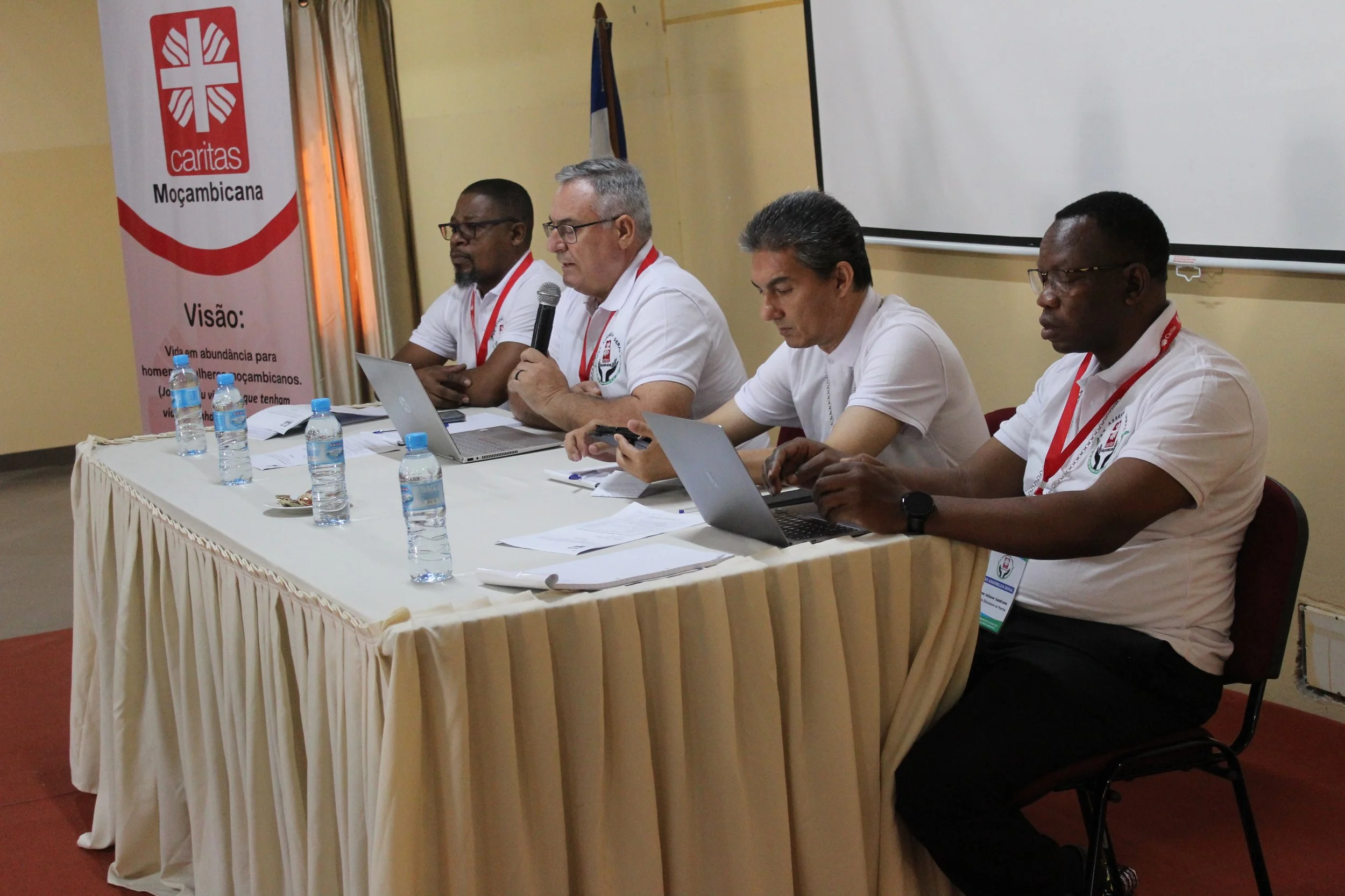 Four men sitting at a long table, participating in a formal meeting or conference. One man is speaking into a microphone, while others are using laptops and mobile phones. There are bottles of water on the table, and a banner with the Caritas logo and text in Portuguese is visible in the background.