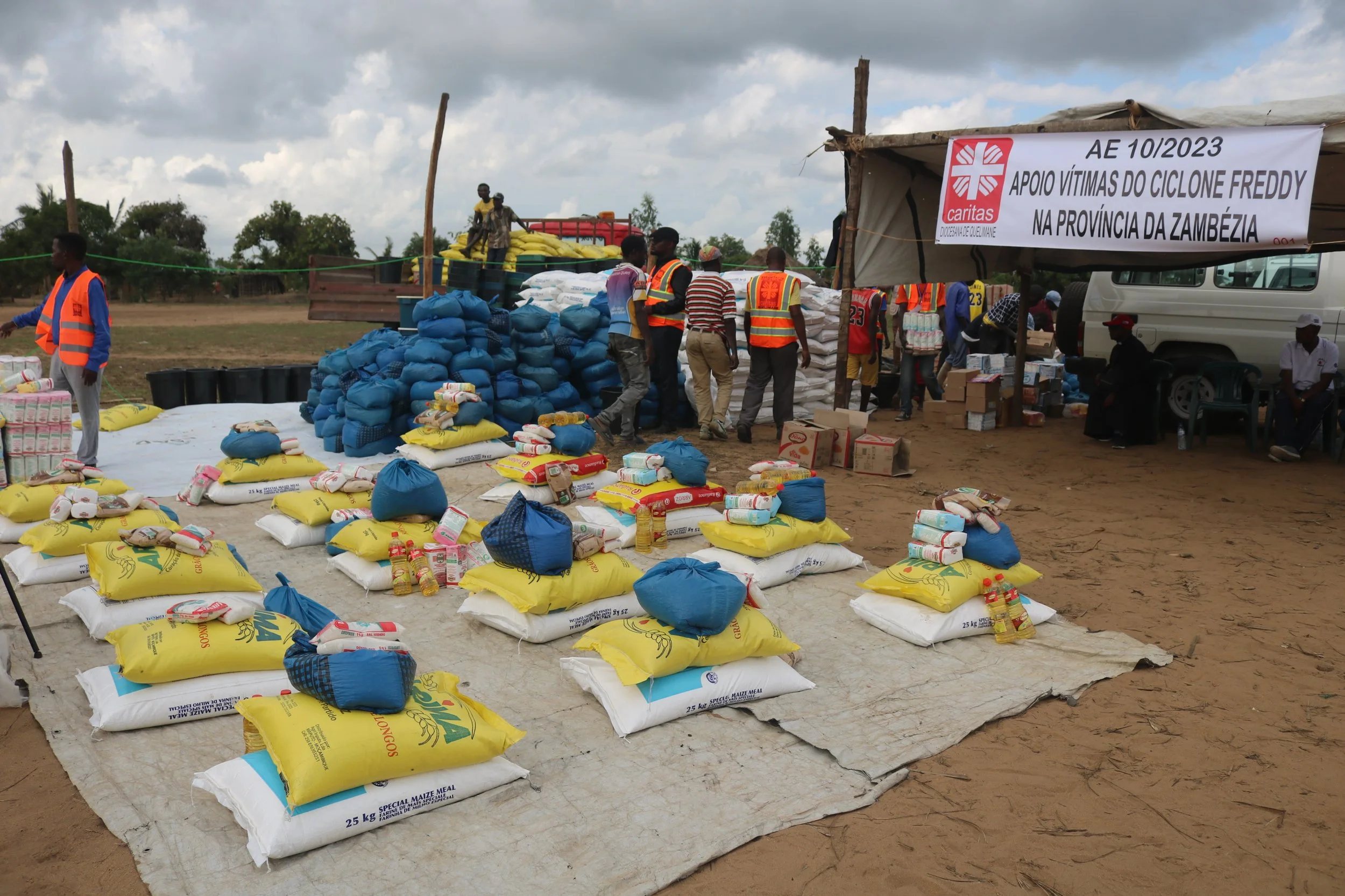 Relief supplies including bags of rice, canned food, and other essentials, are laid out on ground in front of a temporary shelter, with volunteers and aid workers organizing and distributing aid in a rural area affected by cyclone Freddy, Zambia.