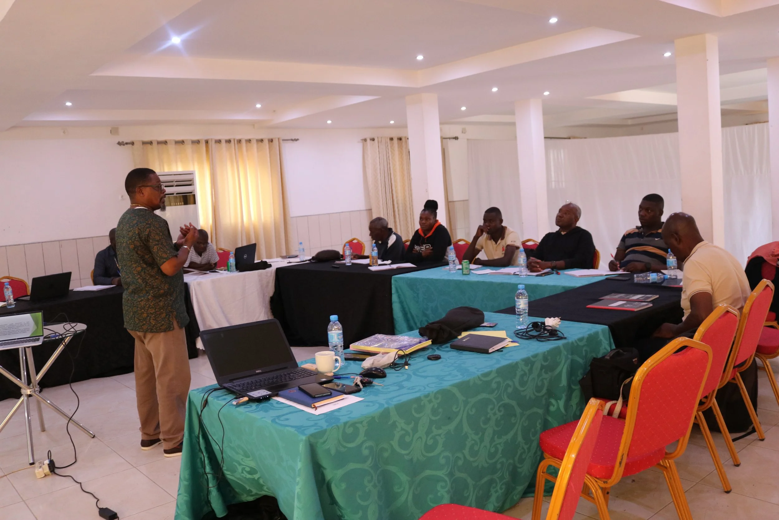 A man in a patterned shirt giving a presentation in front of a seated group in a conference room with tables, laptops, water bottles, and notebooks.