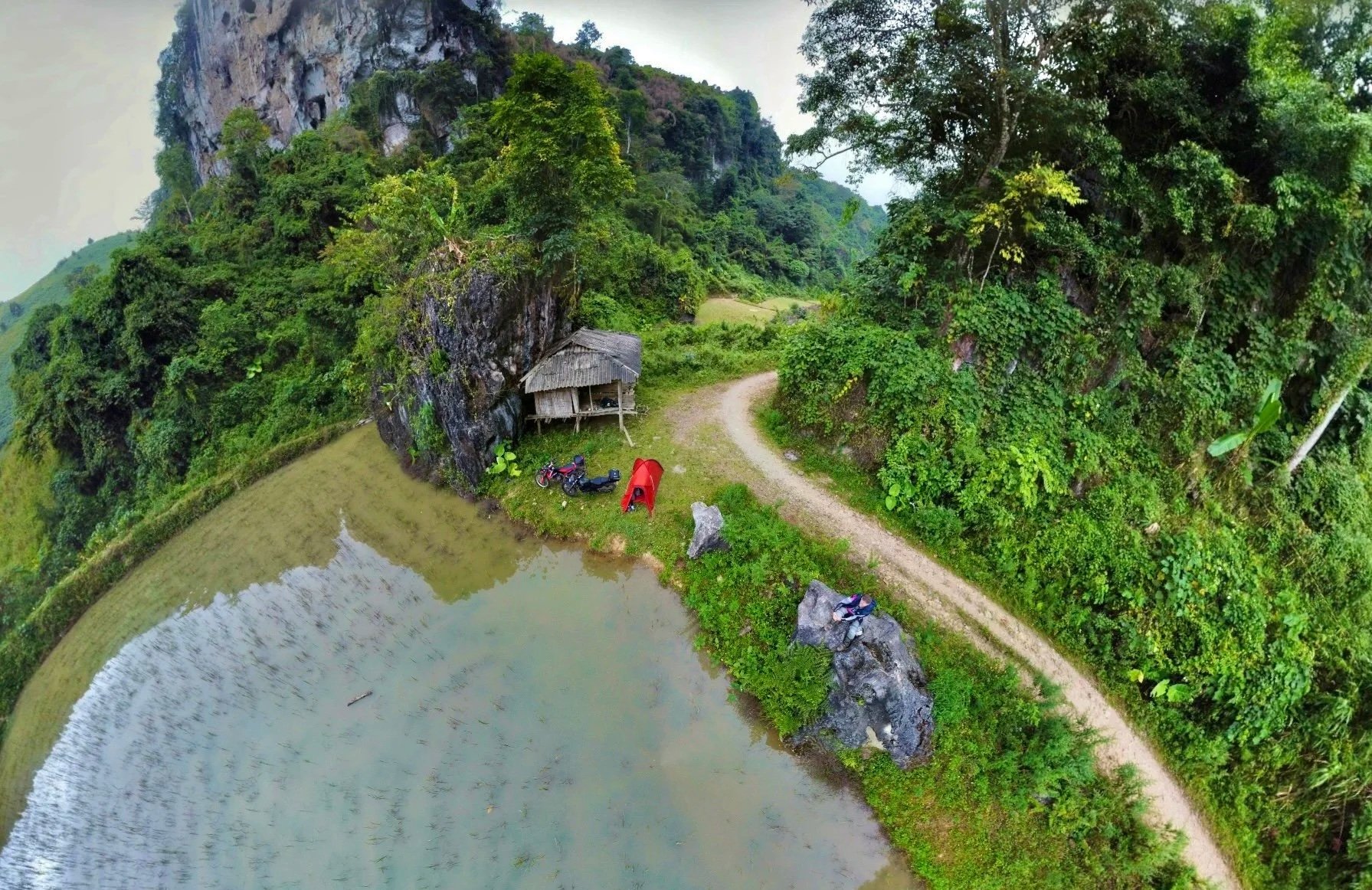 A scenic outdoor area near a small pond with a dirt path, surrounded by lush green trees and bushes. There's a small wooden hut, a red tent, and two motorcycles parked nearby.
