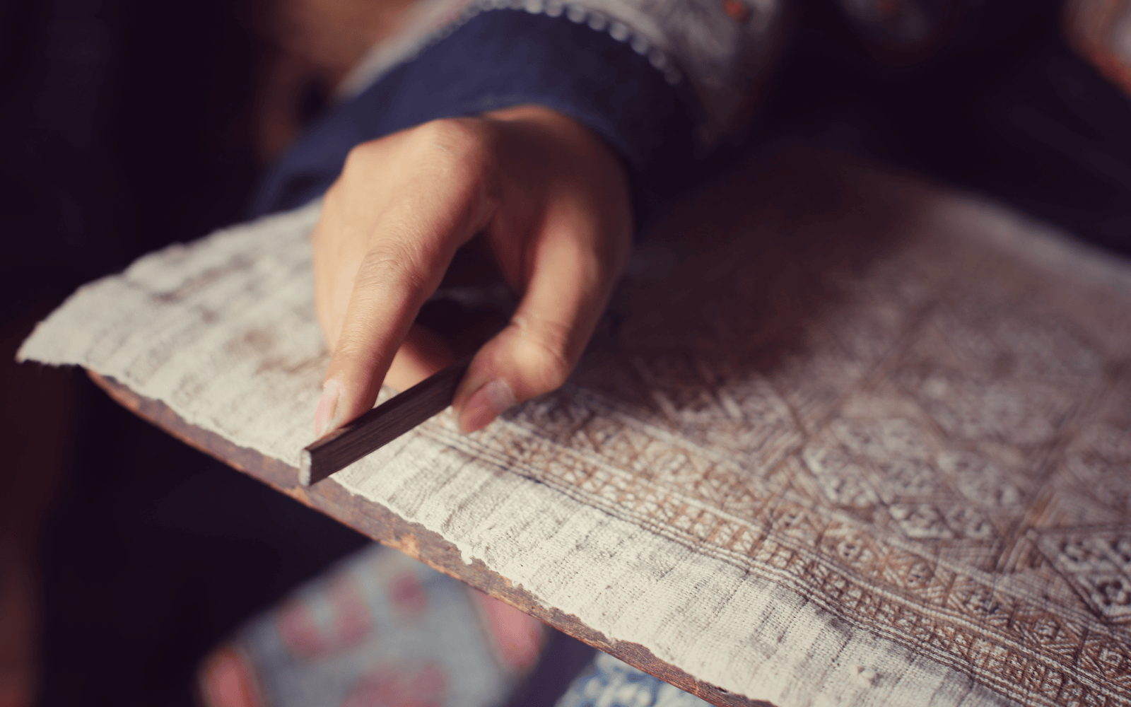 Close-up of hand using a traditional tool to apply wax lines onto fabric, a key step in Hmong batik textile making.