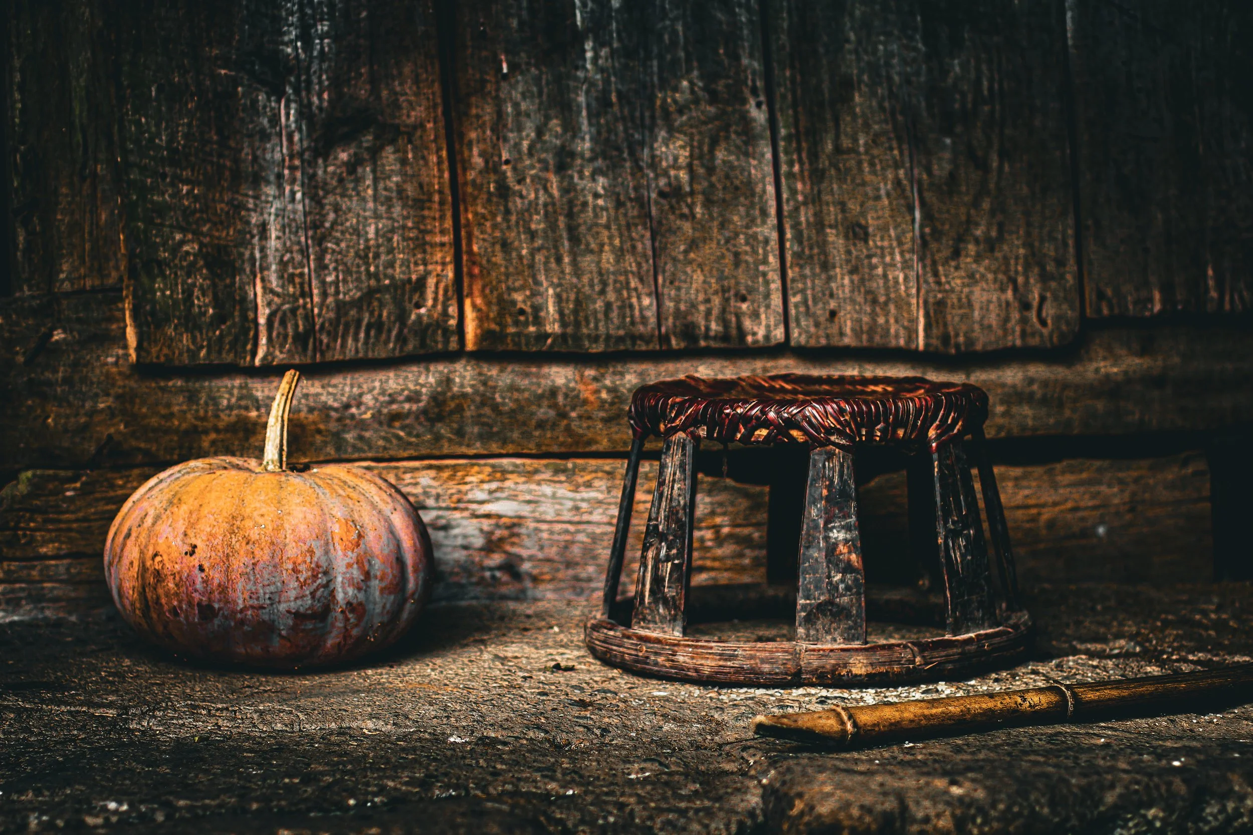 Small woven stool beside an orange pumpkin on an earthen floor against the wooden wall of a Hmong home.