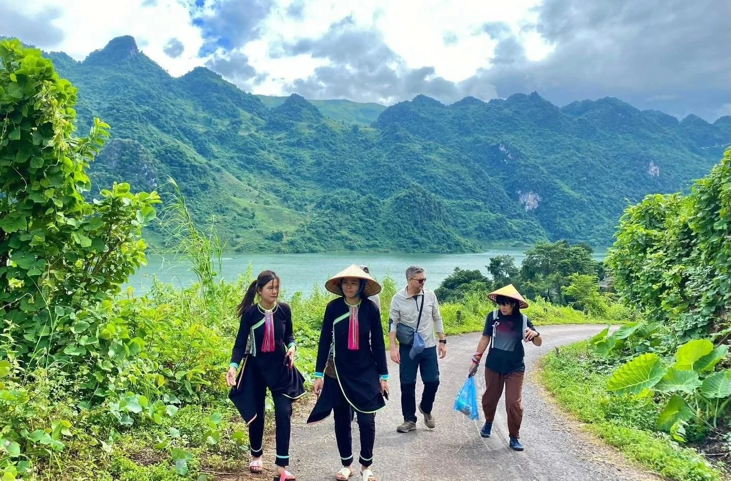 Travellers walking along a rural backroad in Dien Bien Phu with local woman, surrounded by mountains and greenery.