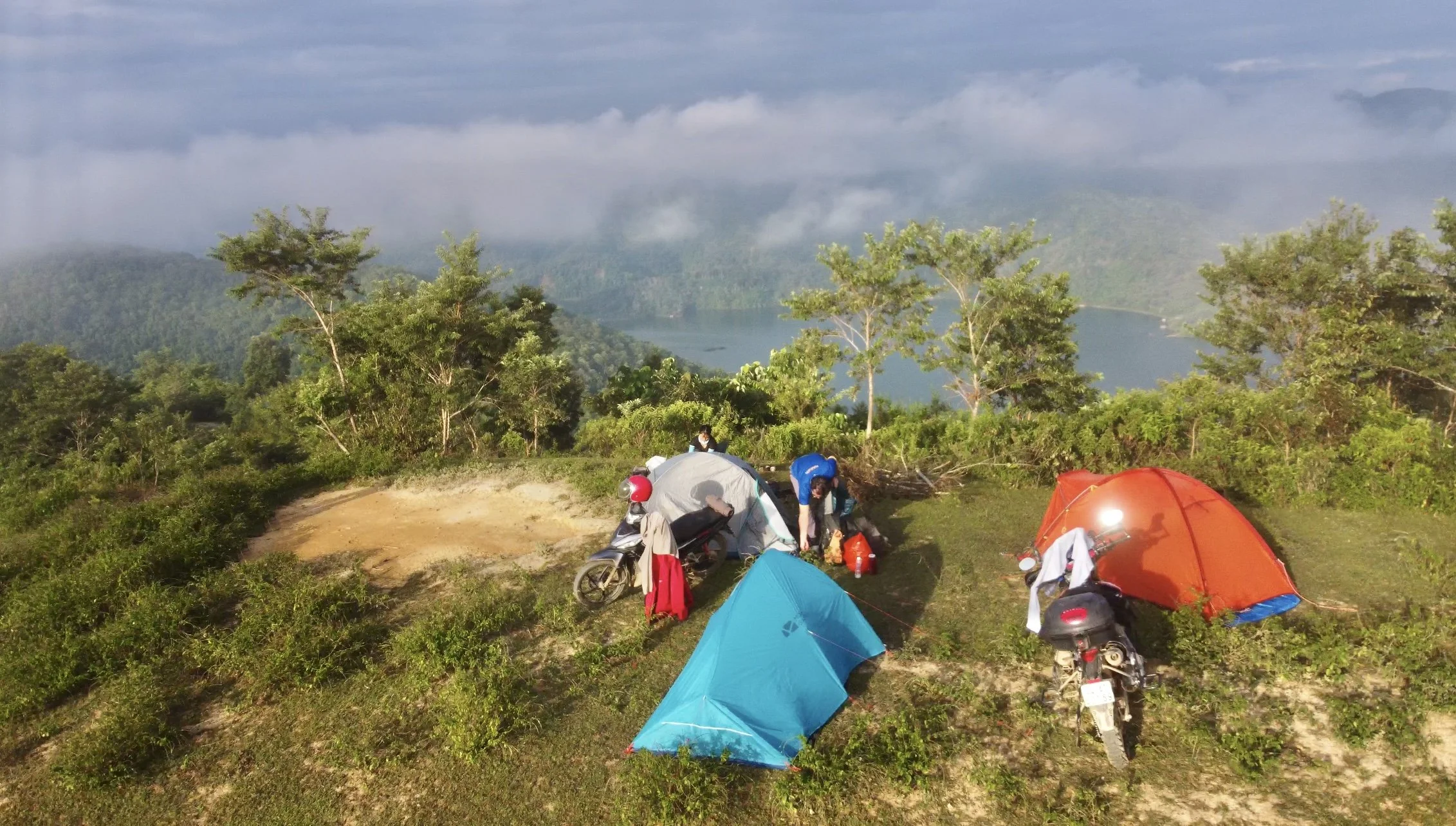 Camping scene with three tents set up on a grassy hill overlooking a lake surrounded by trees and mountains, with two motorbikes and people preparing gear.