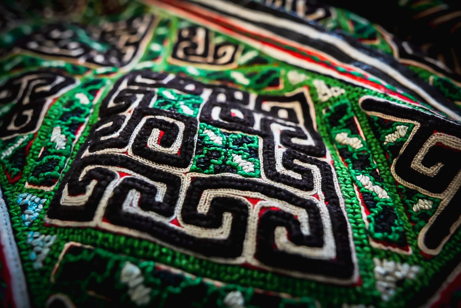 Close-up of Black Hmong embroidery with green stitching, white outlines and black geometric patterns revealing red cloth beneath.