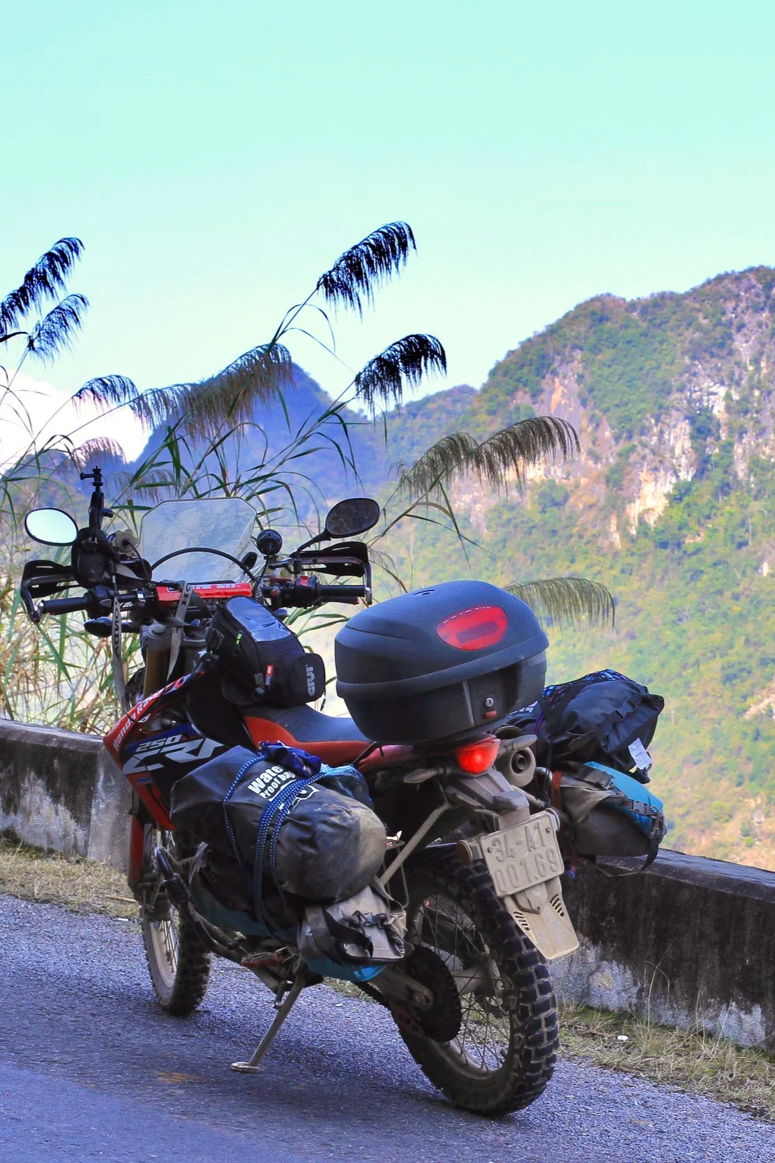 Off-road motorcycle parked on the side of a mountain road, with tall grass and mountains in the background.