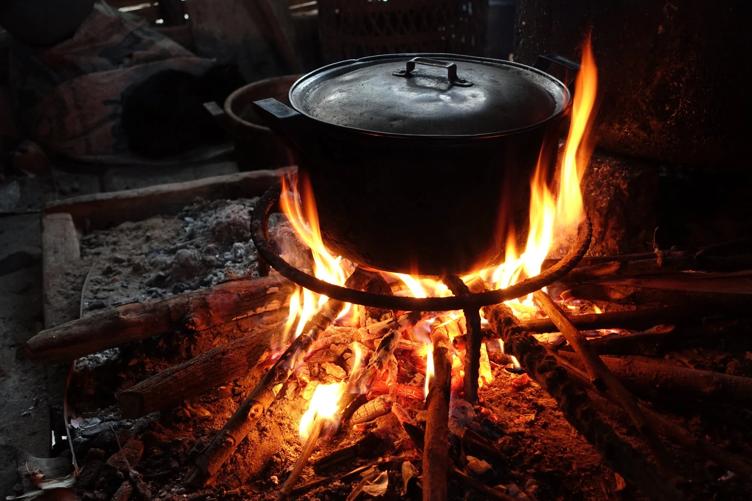 A black cooking pot with a metal lid rests on an iron stand above a bright wood fire.