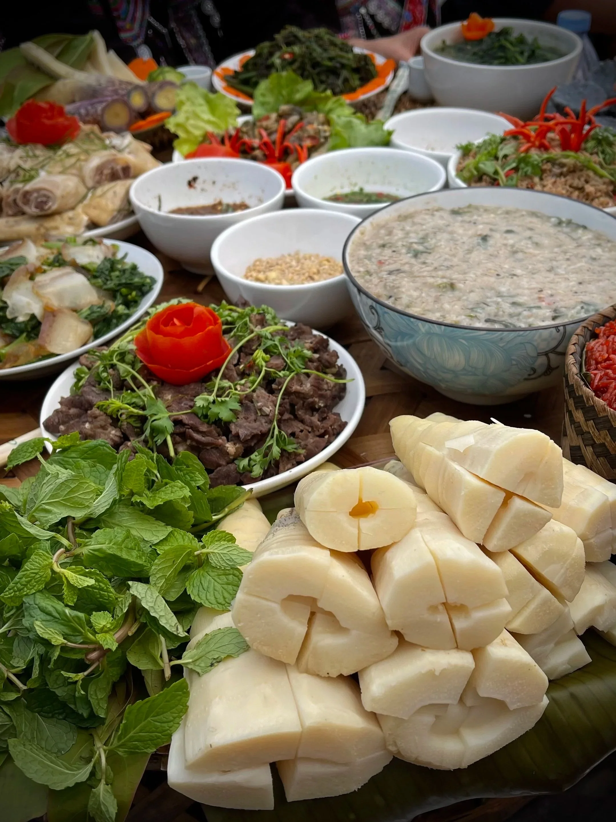 A table filled with Lao festival dishes made from locally farmed and foraged ingredients, including herbs, river plants, and prepared meats, shared during Bun Vốc Nặm celebrations.