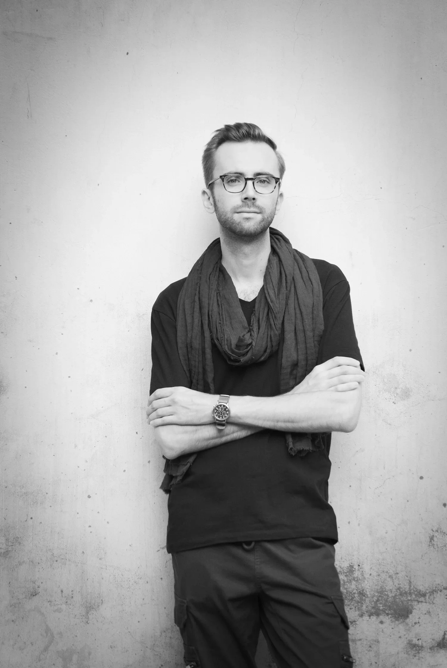 A young man with glasses, a beard, and a scarf standing against a plain wall, arms crossed.