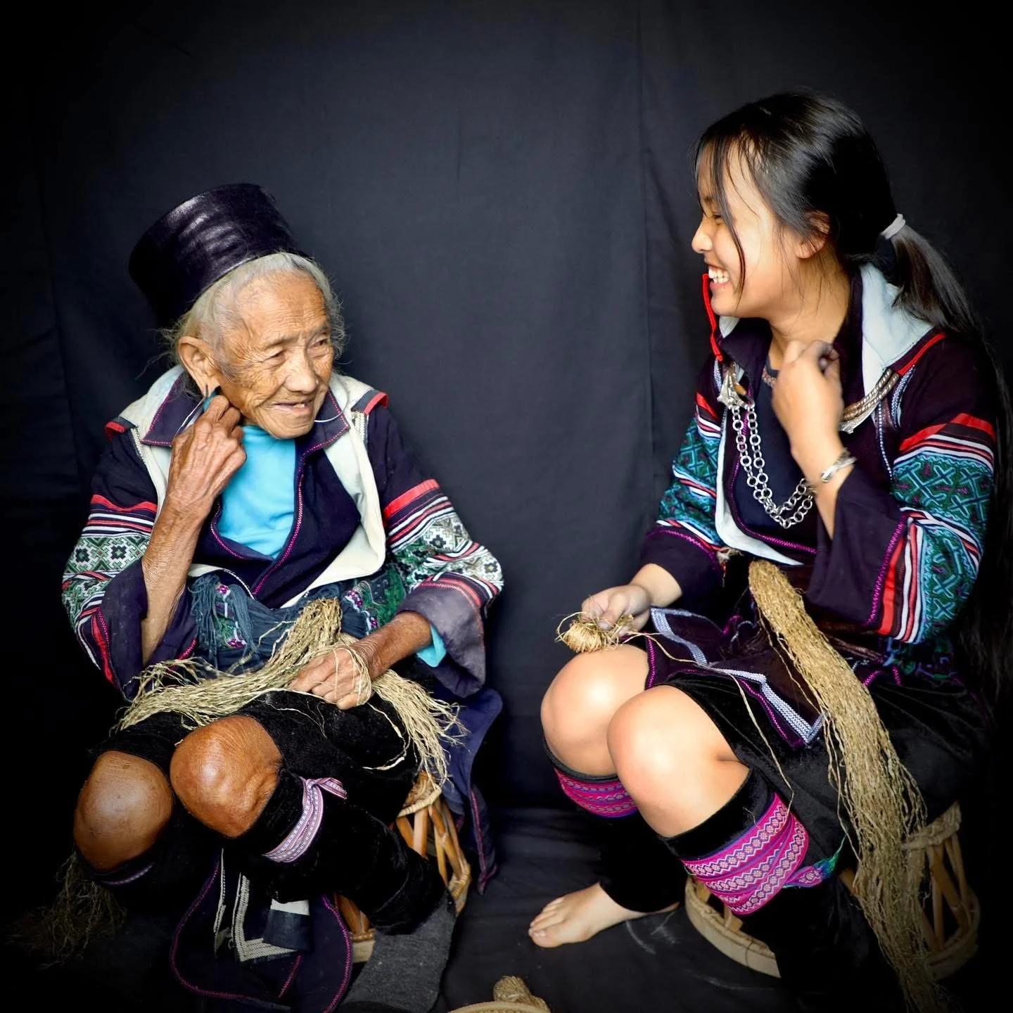 Elderly Hmong grandmother May sitting and talking with a younger woman while holding woven fibers.