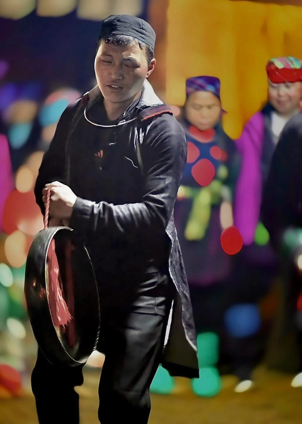 Young Hmong shaman beats a bronze gong while villagers in bright clothing watch during a New Year ceremony.