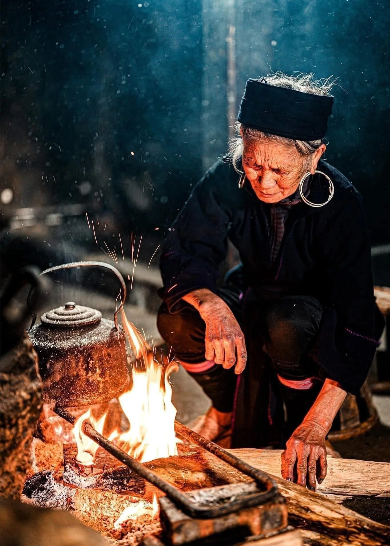An older Hmong woman wearing large hoop earrings tends a wood fire beside a soot-darkened kettle.