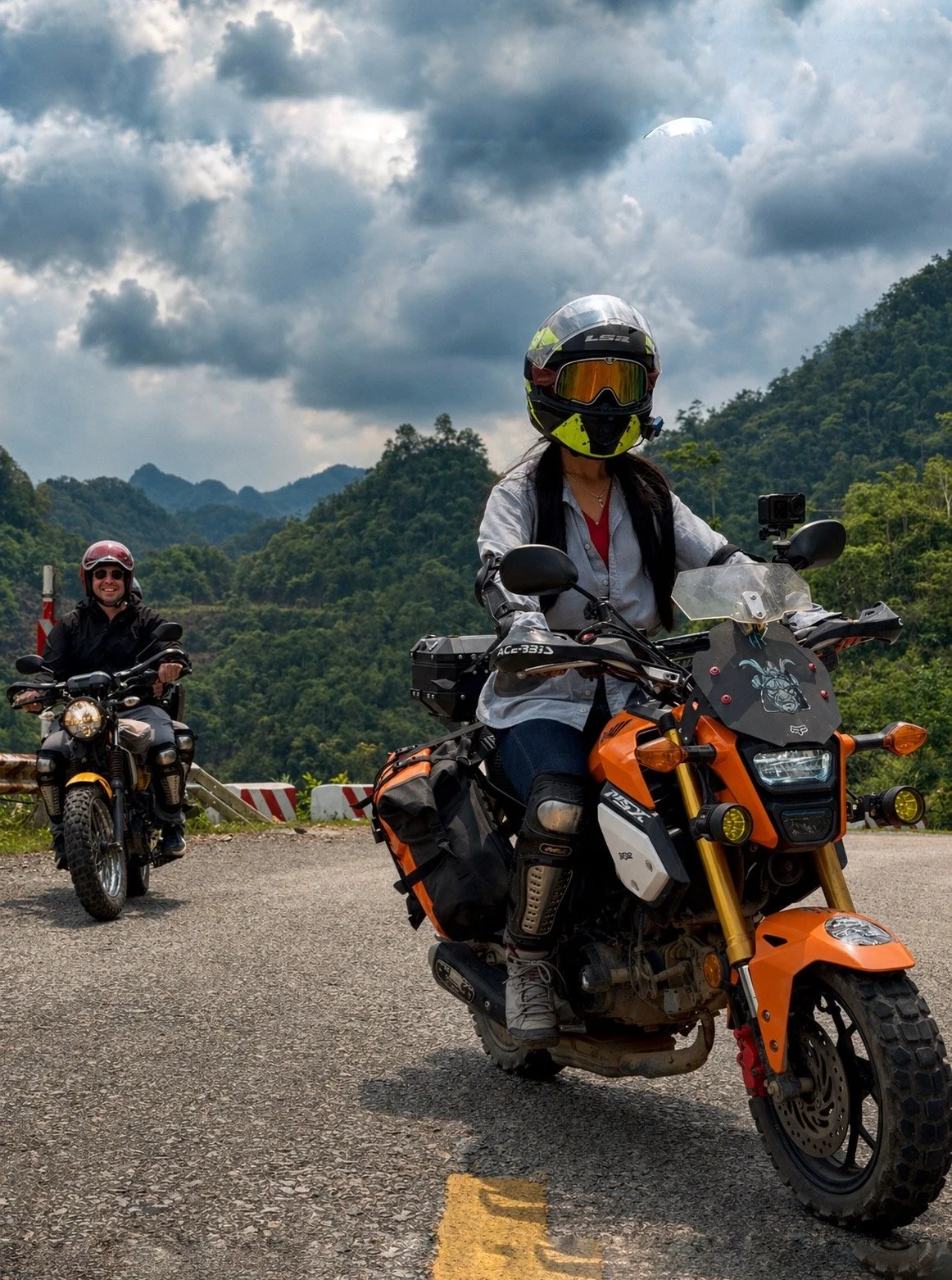 Two riders traveling along a winding mountain road with lush green hills and cloudy skies.