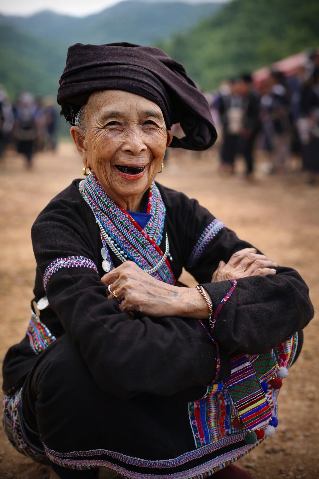 An elderly Lao woman seated with arms folded, dressed in richly embroidered traditional clothing, her expression open and joyful during the spring festival in northern Vietnam.