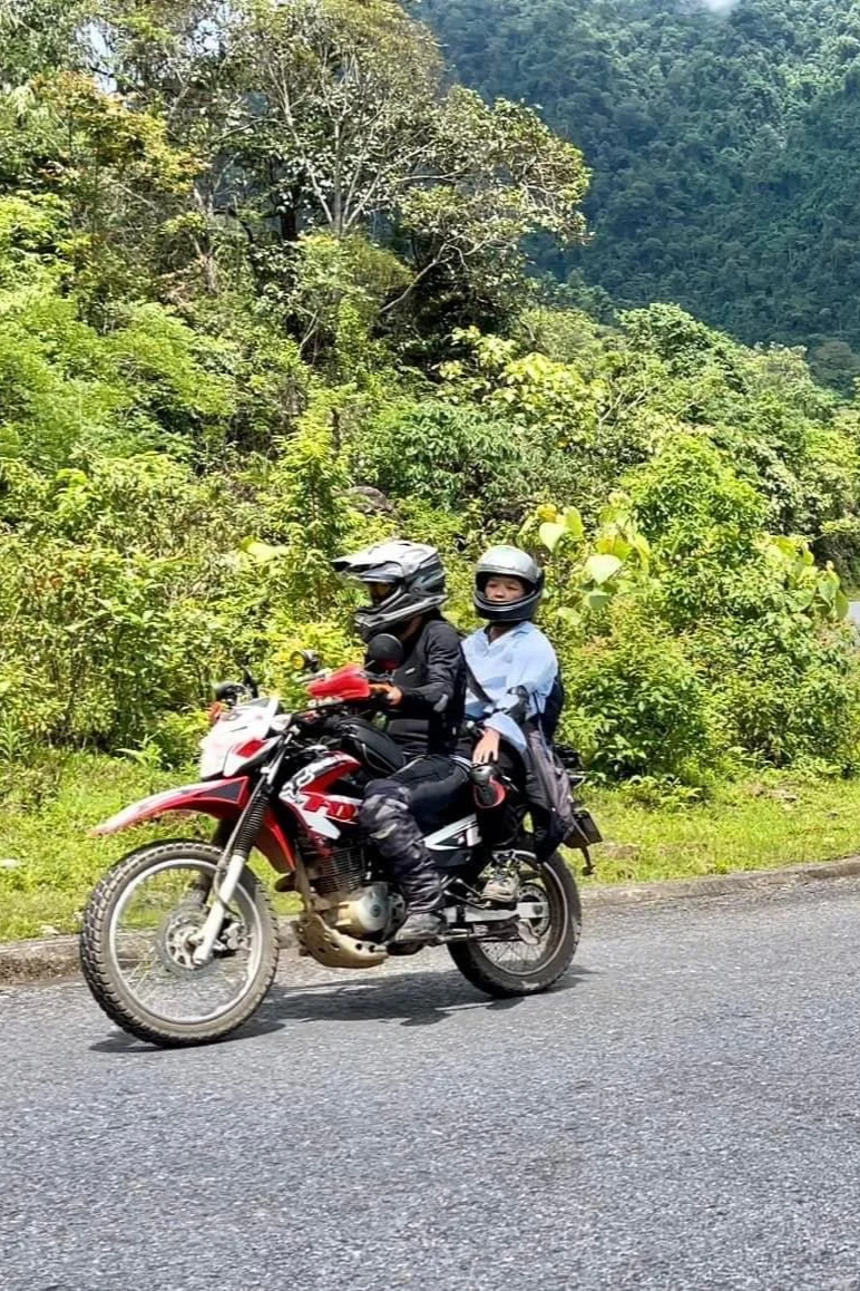 Two people riding a motorcycle on a paved road surrounded by lush green trees and hills.