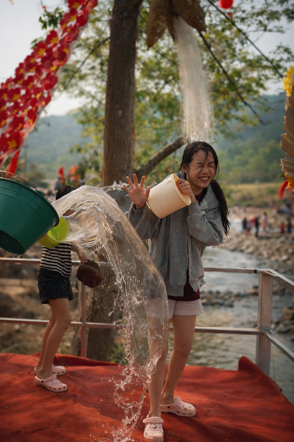 Young Lao girls laughing as water is poured over them from buckets during the Bun Vốc Nặm festival, capturing a moment of surprise, joy, and shared celebration by the riverside.