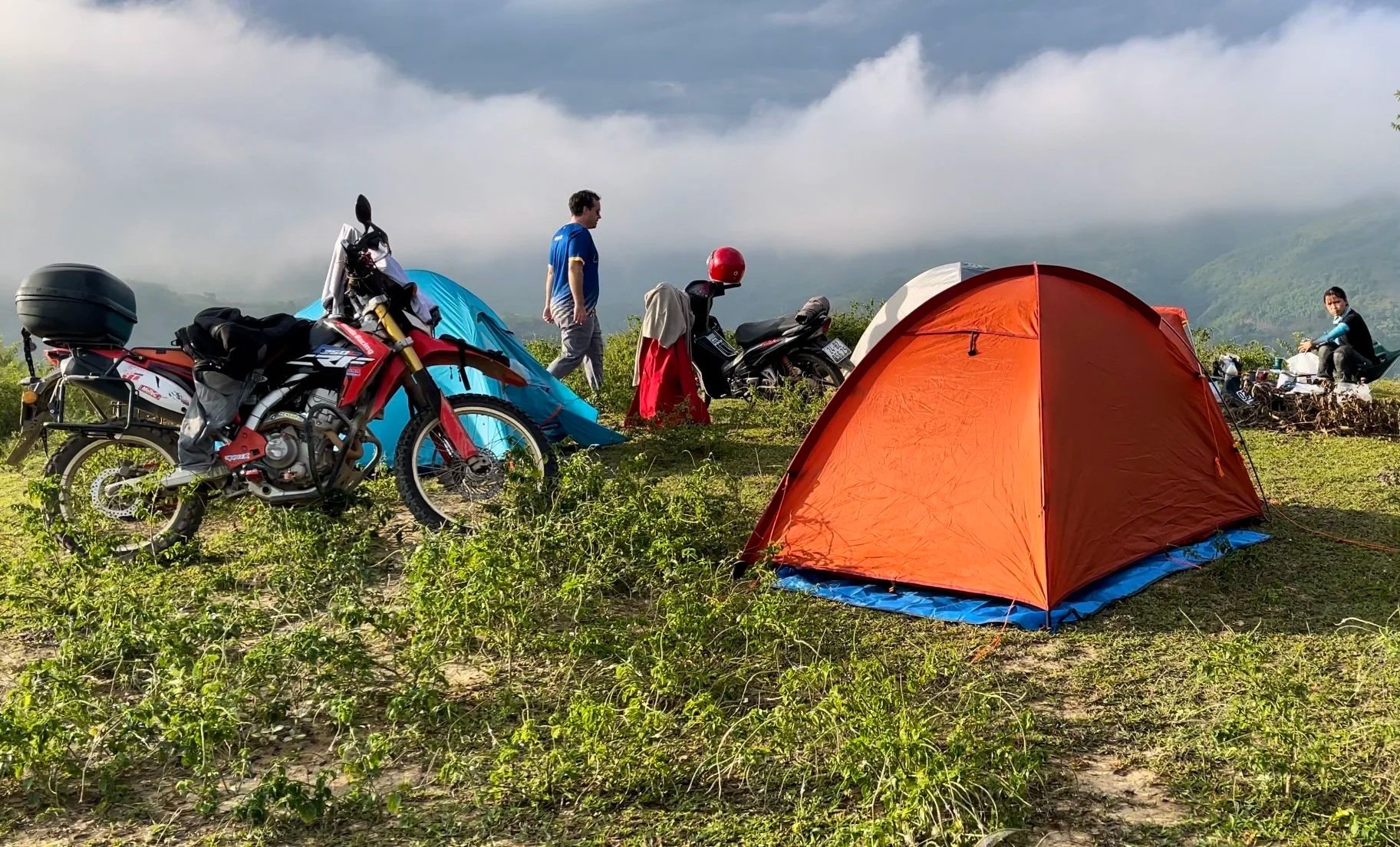 Camping tents, motorcycles, and people on a grassy hillside with mountains and cloudy sky in the background.