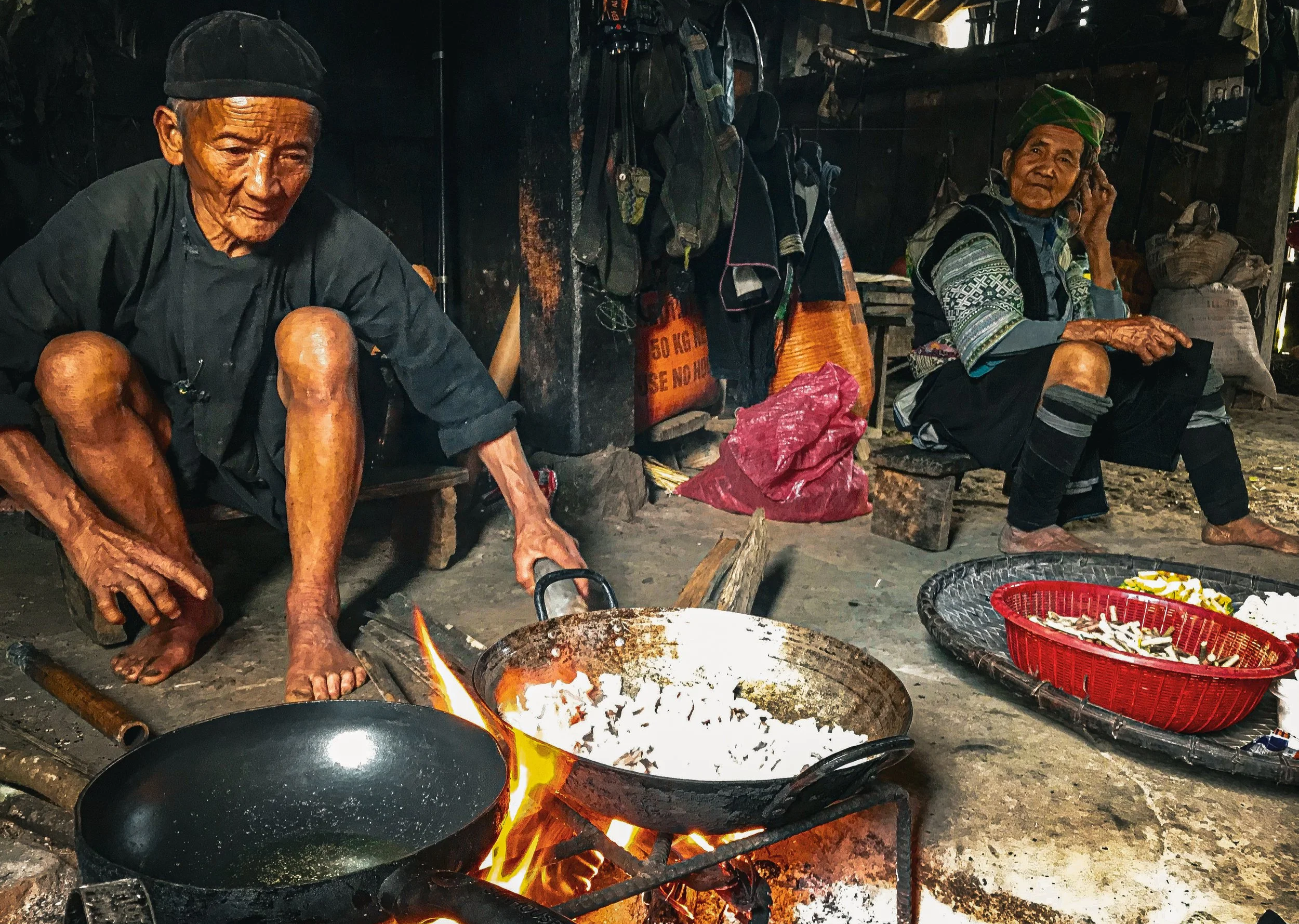An older man tending a wok over an open hearth while an older woman sits nearby inside a Hmong home.
