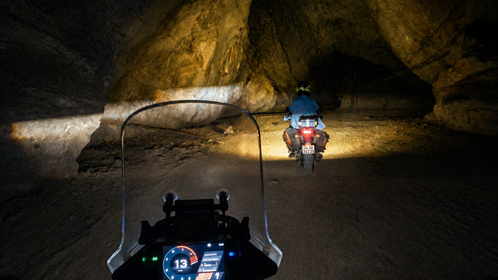 Motorcyclist riding through a dimly lit cave tunnel with headlights illuminating rocky walls.