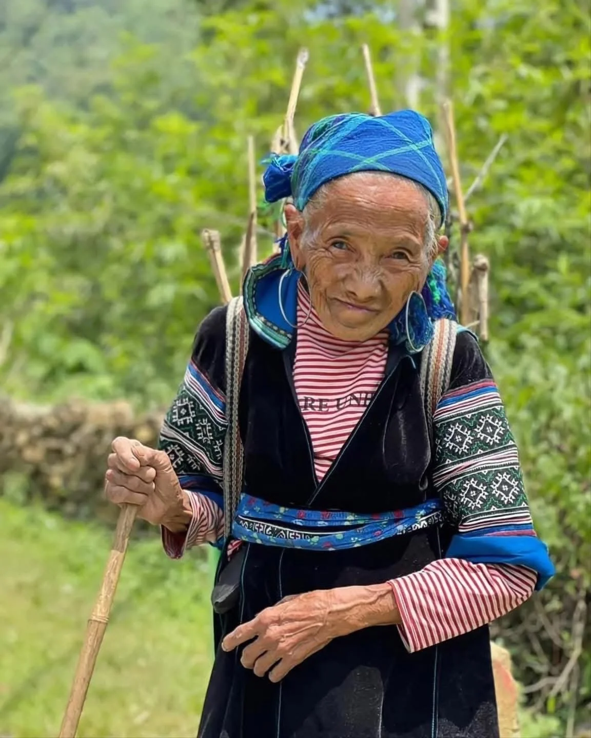 Elderly Hmong grandmother May standing outdoors with a walking stick, wearing traditional clothing in a green mountain landscape.