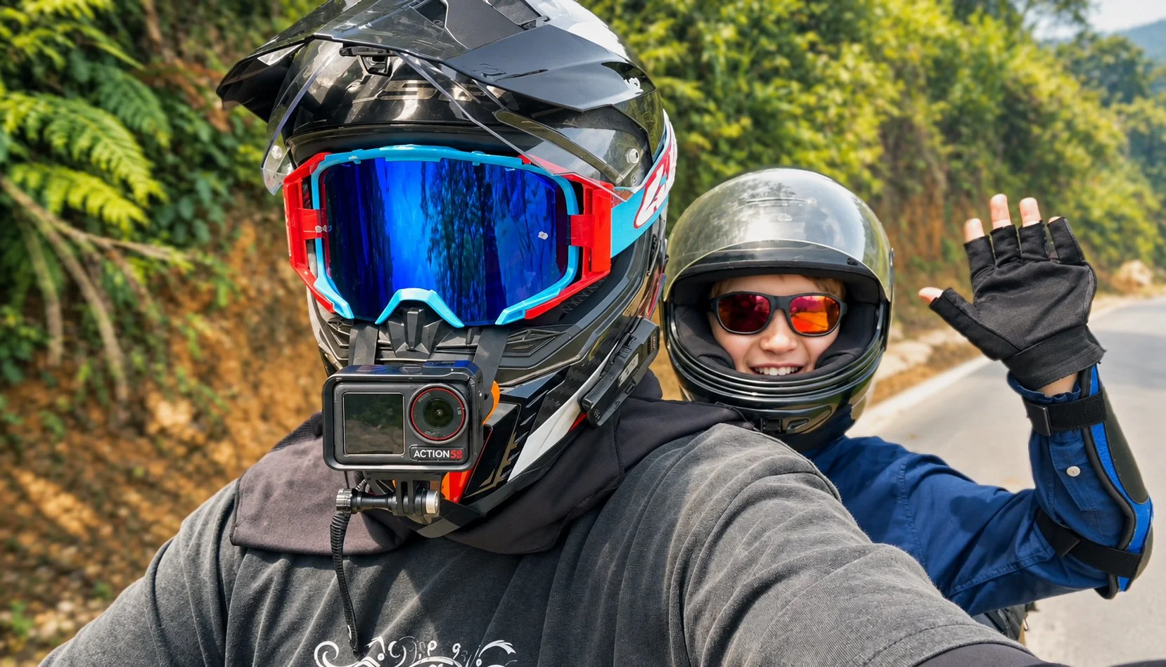 Two people wearing motorcycle helmets and gear taking a selfie on a scenic road surrounded by trees. One is waving and both are smiling.